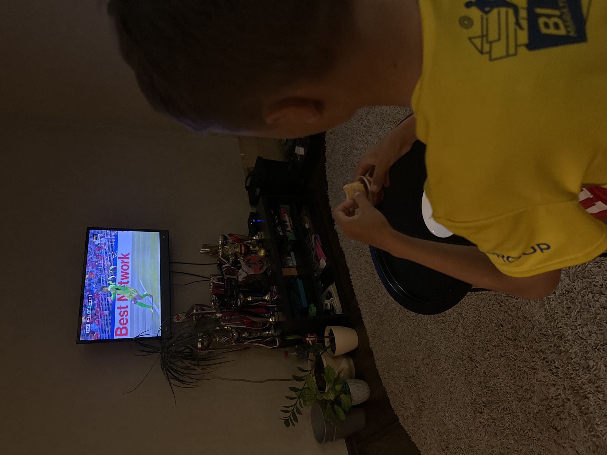 A boy in a yellow t-shirt with a graphic on the back, likely a logo from a marathon or sporting event, is sitting in front of a television in a dimly lit room, eating from a small red container with a chip. The television displays a soccer match with the score at 0-0, and a banner that reads "Best Network." The room appears to be a living space, with a black entertainment center behind the TV filled with trophies, potted plants, and various containers. A plush rug covers the floor. The scene suggests a casual evening at home, possibly in Chaikovsky, Russia, given the context.