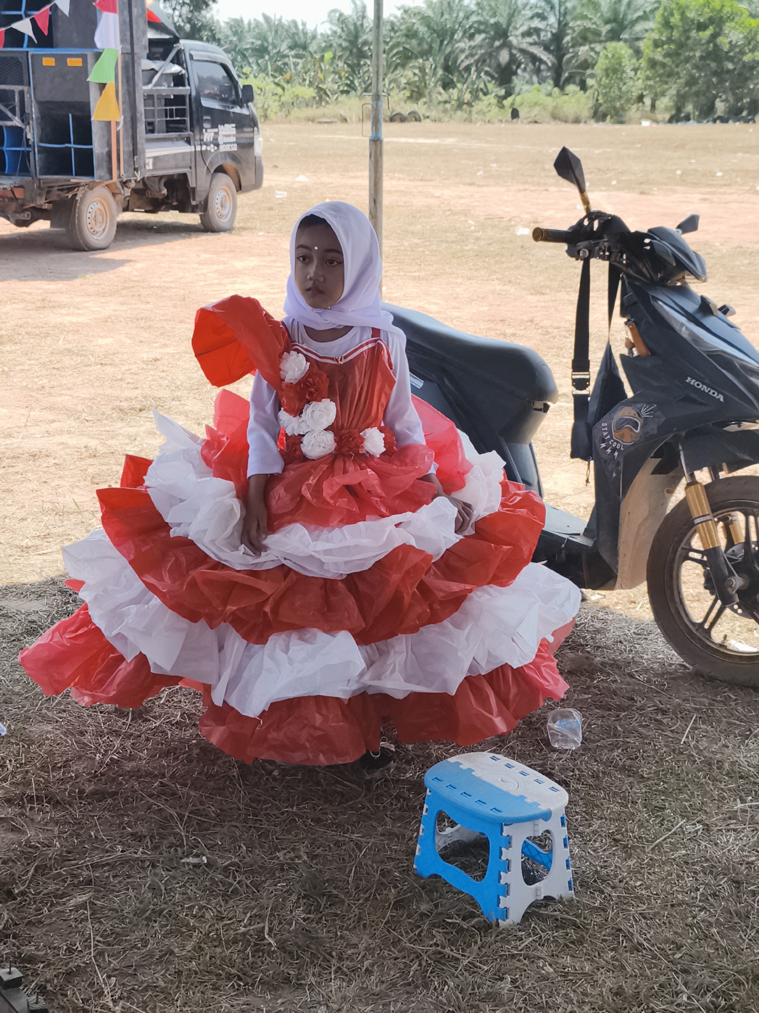 In Pelanjau Jaya, Indonesia, a young girl wearing a red and white ruffled dress and a white hijab sits on the ground, looking off to the side. The dress features a voluminous skirt made of layered red and white ruffles, adorned with white floral accents on the bodice. Beside her, a small blue and white folding stool sits on the dry, grassy ground. In the background, a black truck with speakers and flags is parked, and a black scooter with gold accents is also visible. The scene appears to be outdoors in a rural or semi-rural setting, with palm trees and vegetation in the distance.