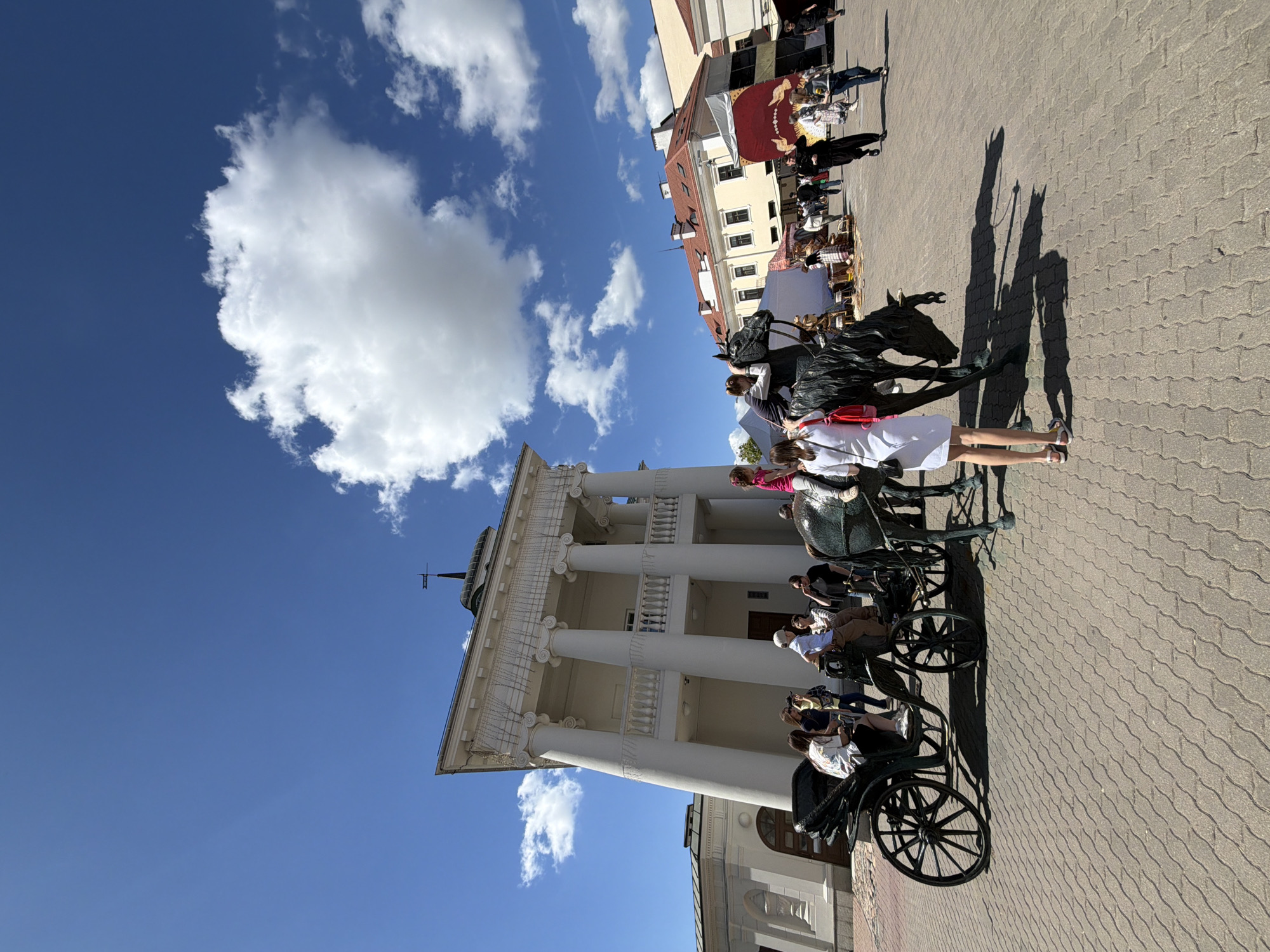 A horizontal outdoor shot captures a scene at the City Hall Square in Minsk, Belarus. The City Hall stands prominently with its white neoclassical facade, tall columns, a balcony, and a cross on its roof. A bright blue sky is partially filled with puffy white clouds. Multiple horse-drawn carriages are parked in front of the building, filled with passengers. A bronze horse statue is situated between the carriages and the building, with people standing next to it, appearing to take photos. In the background, stalls of a street fair are visible, occupied by people. The ground is paved with gray bricks. The overall lighting indicates a sunny day.