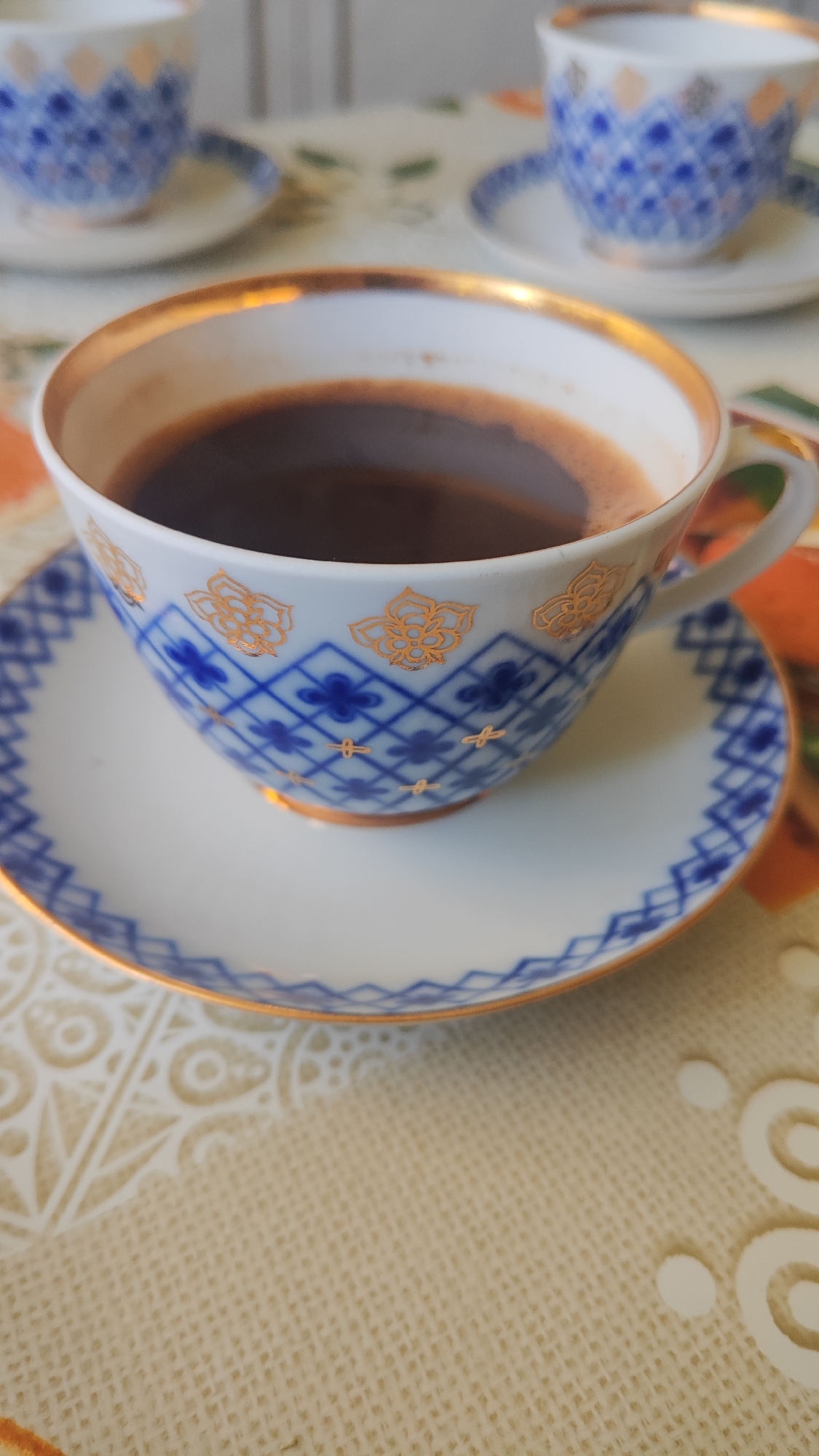 The image displays a close-up of a white porcelain teacup filled with dark liquid, likely coffee. The teacup has a gold rim and is adorned with a blue and gold intricate pattern of diamonds and floral motifs. It rests on a matching saucer with a similar blue and gold border. In the background, out of focus, are two more teacups and saucers, also featuring the same blue and gold pattern, suggesting a set. The cups are placed on a textured tablecloth with a subtle beige color and a cream-colored lace-like pattern. The lighting is natural and soft, highlighting the gleam of the gold trim. The scene appears to be set indoors, possibly in a domestic setting or a cafe. While the image is undated and does not contain explicit markers of location, the provided context indicates it is in Dagomys, Russia. There are no people visible in the image.