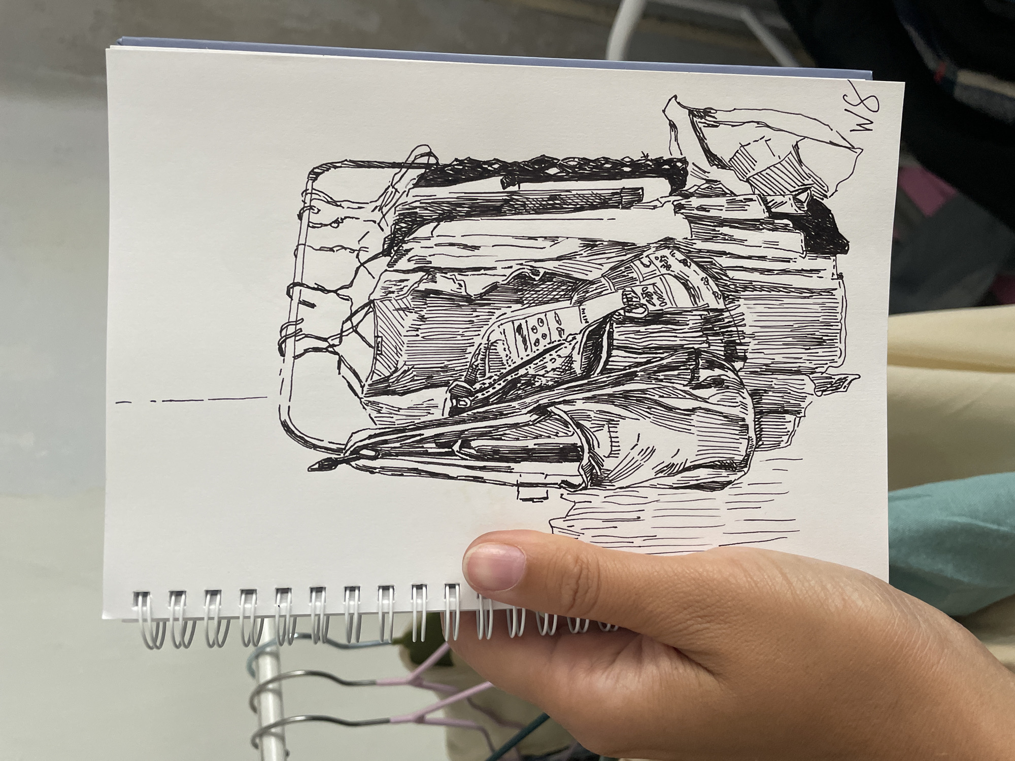 The image shows a hand holding a spiral-bound sketchbook. The sketchbook contains a black ink drawing of a clothing rack with several garments and a bag hanging on it. The drawing is done in a sketchy, realistic style with heavy use of hatching to create shadows and texture. The background of the drawing is white, indicating it is on paper. Behind the sketchbook, the actual clothing rack with real clothes and hangers is visible, suggesting the drawing is a study of the real items. The overall setting appears to be indoors, possibly a bedroom or dressing area, with hints of other objects and fabrics in the background. The lighting is natural and somewhat diffused.