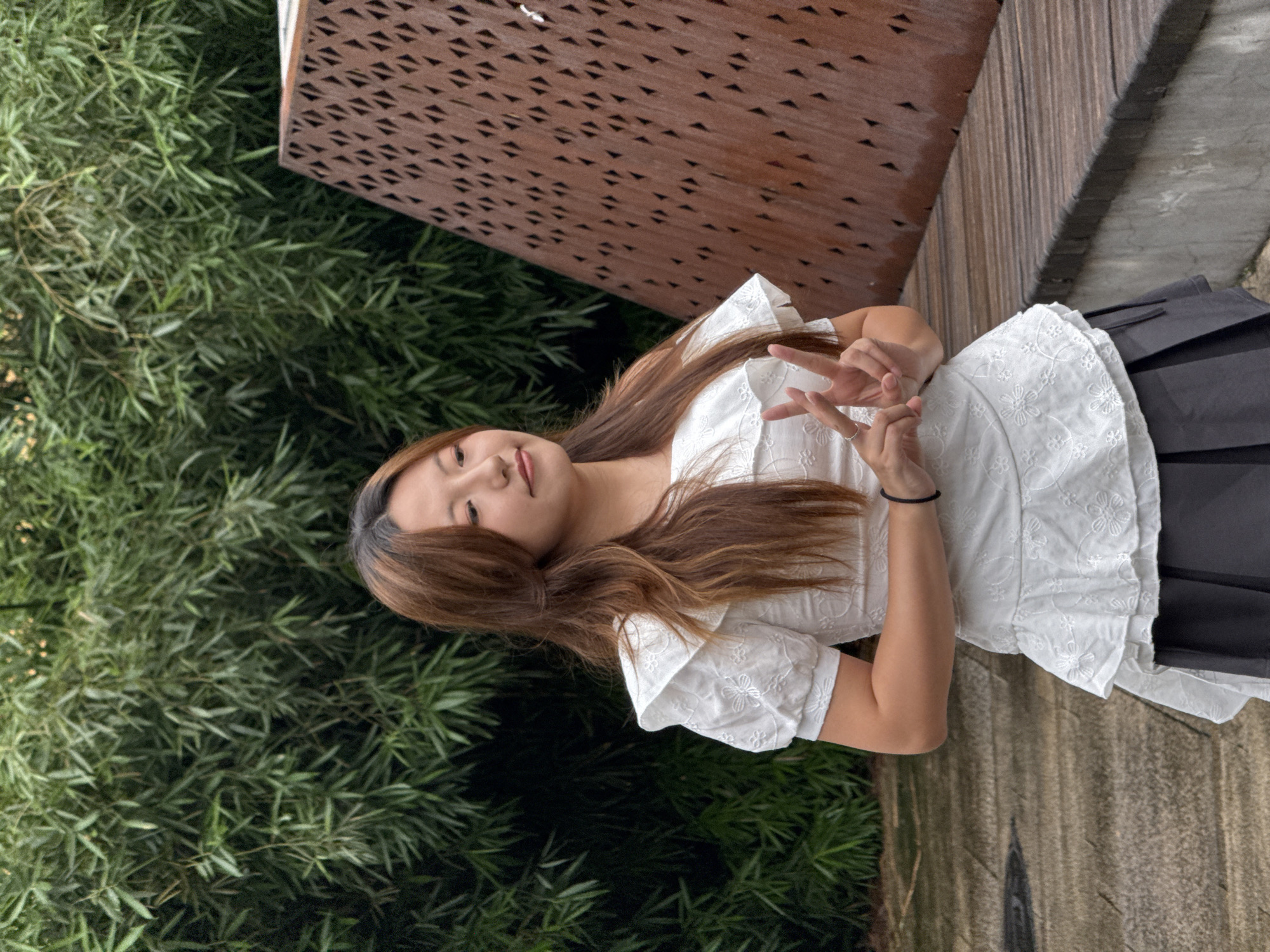A young Asian woman with long, brown hair is standing outdoors in front of a bamboo forest and a wooden structure. She is wearing a white, off-the-shoulder top with ruffled details and floral embroidery, and a dark gray pleated skirt. Her left hand is raised, forming a peace sign. The background consists of dense green bamboo foliage and a rustic, angular wooden bench and partition. The ground beneath her appears to be a wet, stone pathway. The scene is consistent with a location in the Yuzhong District, China.