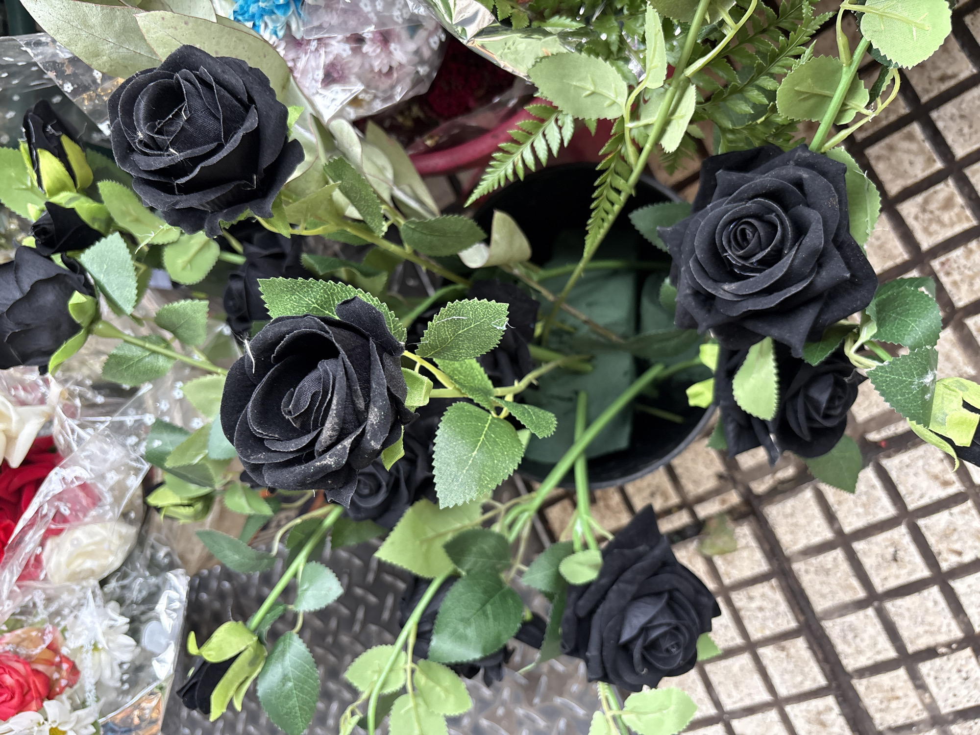 An overhead shot of several bunches of artificial flowers. Dominating the view are black roses with green leaves. There are several roses, and they appear to be in a bucket or container. Adjacent to the black roses, partially visible, are other bouquets with white, red, and blue flowers wrapped in clear cellophane. In the upper right corner, fern-like greenery is visible. The surface below is a tiled, grid-like pattern. The image was likely taken in Buenos Aires, Argentina.