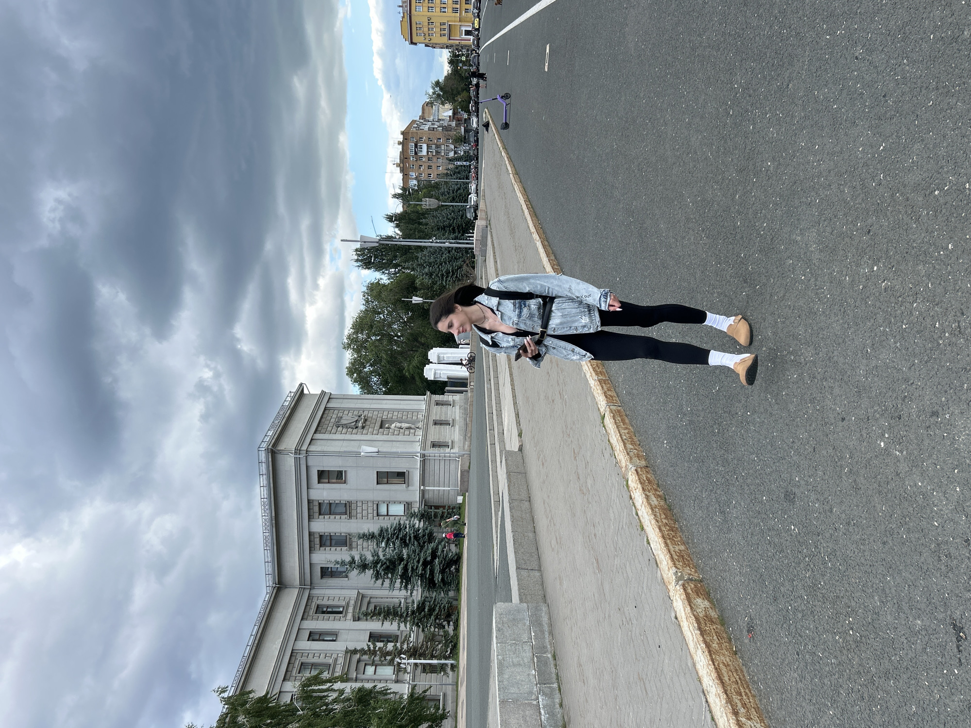 This image shows a young woman standing on a street in Novokuybyshevsk, Russia. She is wearing a denim jacket, a light-colored top, black leggings, white socks, and tan shoes. She is holding a phone in her hand and has a bag slung across her shoulder. The woman is looking down at her phone.
The scene is an urban street with a sidewalk and a road. A large, imposing building with classical architecture and trees in front of it is visible on the left side of the image. The sky is cloudy, suggesting overcast weather. In the background, there are more buildings and parked cars. A purple scooter is visible on the road. The time of day appears to be daytime, likely afternoon, given the lighting conditions. There is no visible text in the image. The woman's posture and the casual attire suggest a relaxed, everyday scene.