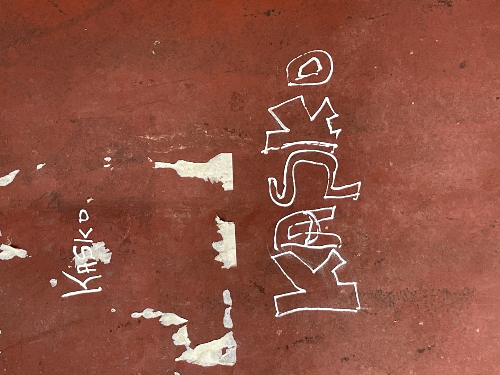 The image depicts a maroon-colored surface with white markings. The word "Kasko" is written in white paint in two locations. One instance is smaller and located on the left side of the image. The other, larger instance is oriented vertically on the right side. There are areas where the maroon surface is chipped or worn, revealing a lighter-colored material underneath. The scene is likely outdoors, and the location is assumed to be Mararaba, Nigeria. No people or other identifiable subjects are visible. The interaction is limited to the markings on the surface.