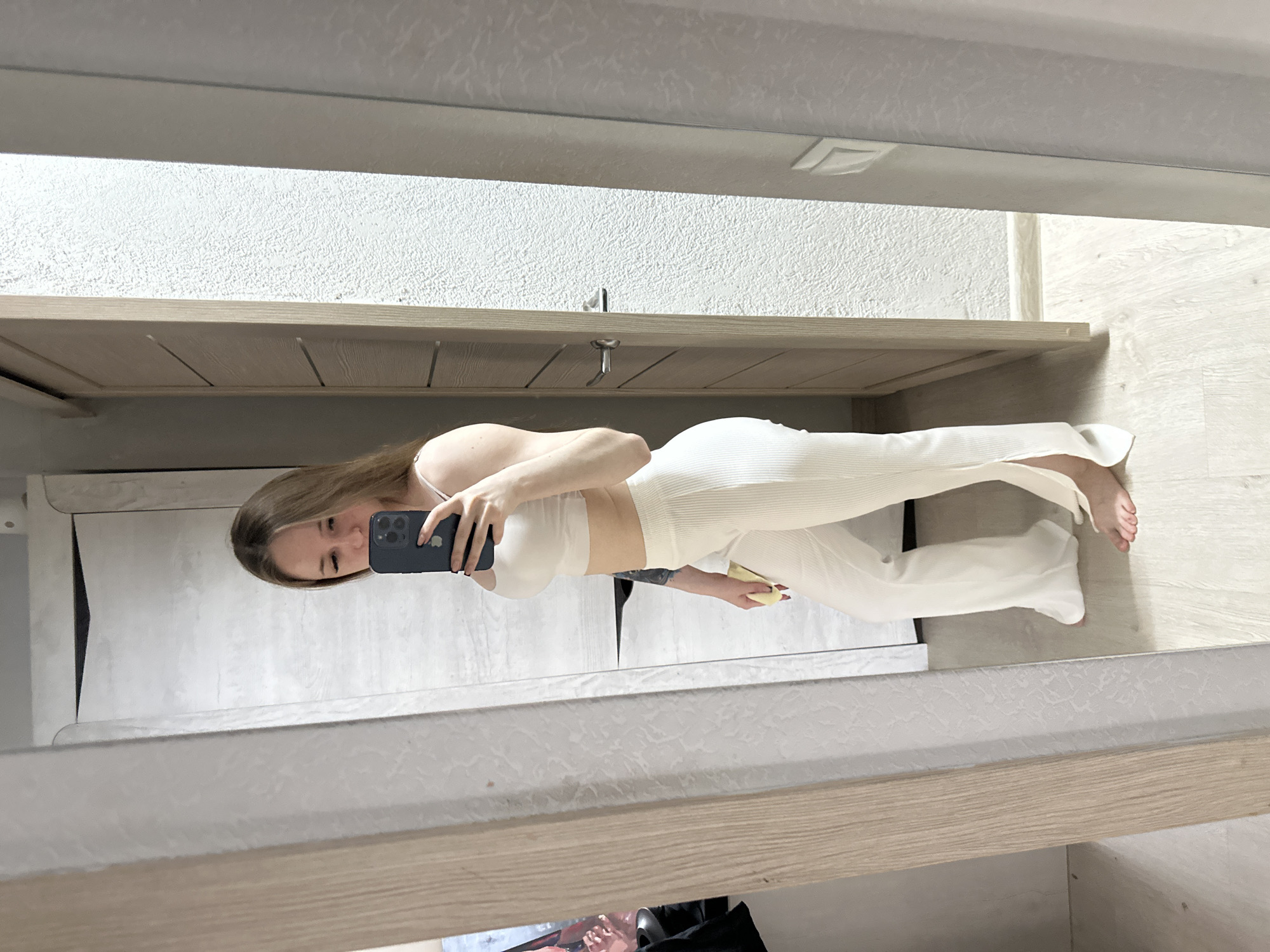 A woman is captured in a full-length mirror selfie, with the image rotated 90 degrees clockwise. She stands with her body slightly turned, holding a dark-colored smartphone, possibly an iPhone, up to her face, looking down at the screen. She has light brown hair that falls over her shoulders and is wearing a matching off-white, ribbed crop top and flared or wide-leg pants. Her fingernails are painted red. In her left hand, she holds a small yellow object. The reflected background shows a light-colored, textured wall above a light wooden cabinet or shelf that features recessed panels and a silver handle. The floor visible in the reflection is a light wood laminate. No text is visible in the image.