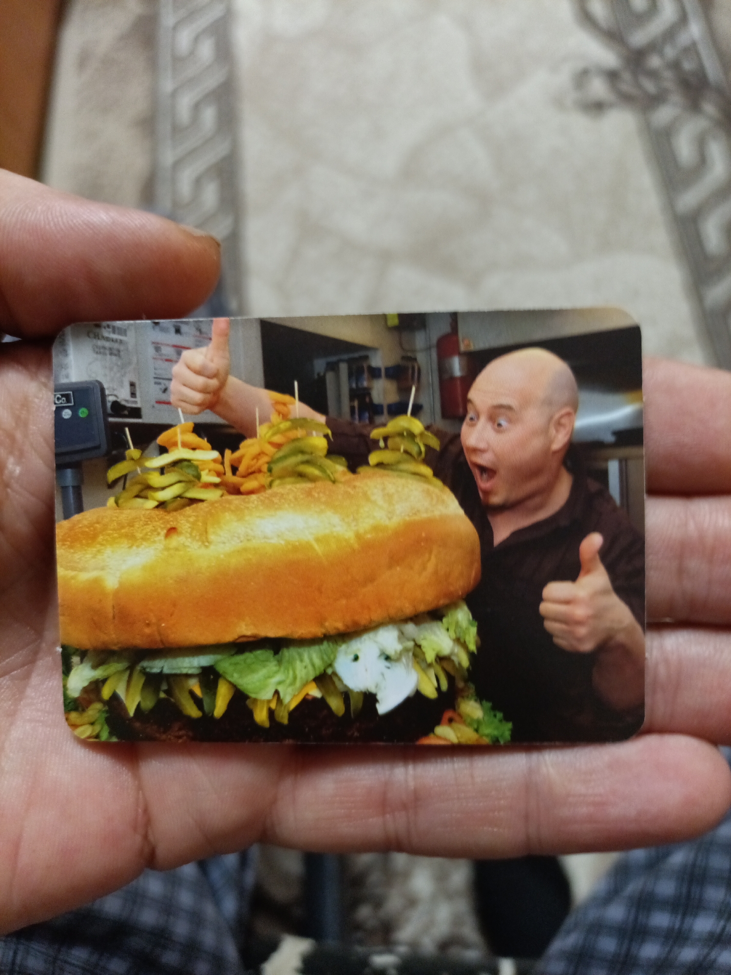 The image shows a postcard-sized photograph being held by a person's hands. The photograph depicts a giant hamburger with pickles and fries on top. A bald man is visible in the background, making a surprised expression and giving a thumbs-up. A scale is also visible on the left side of the photograph. The background of the postcard shows a busy kitchen environment. The postcard itself is lying on a surface with a textured pattern, possibly a blanket or upholstery. There is no information in the image to suggest the location in Nyagan, Russia.