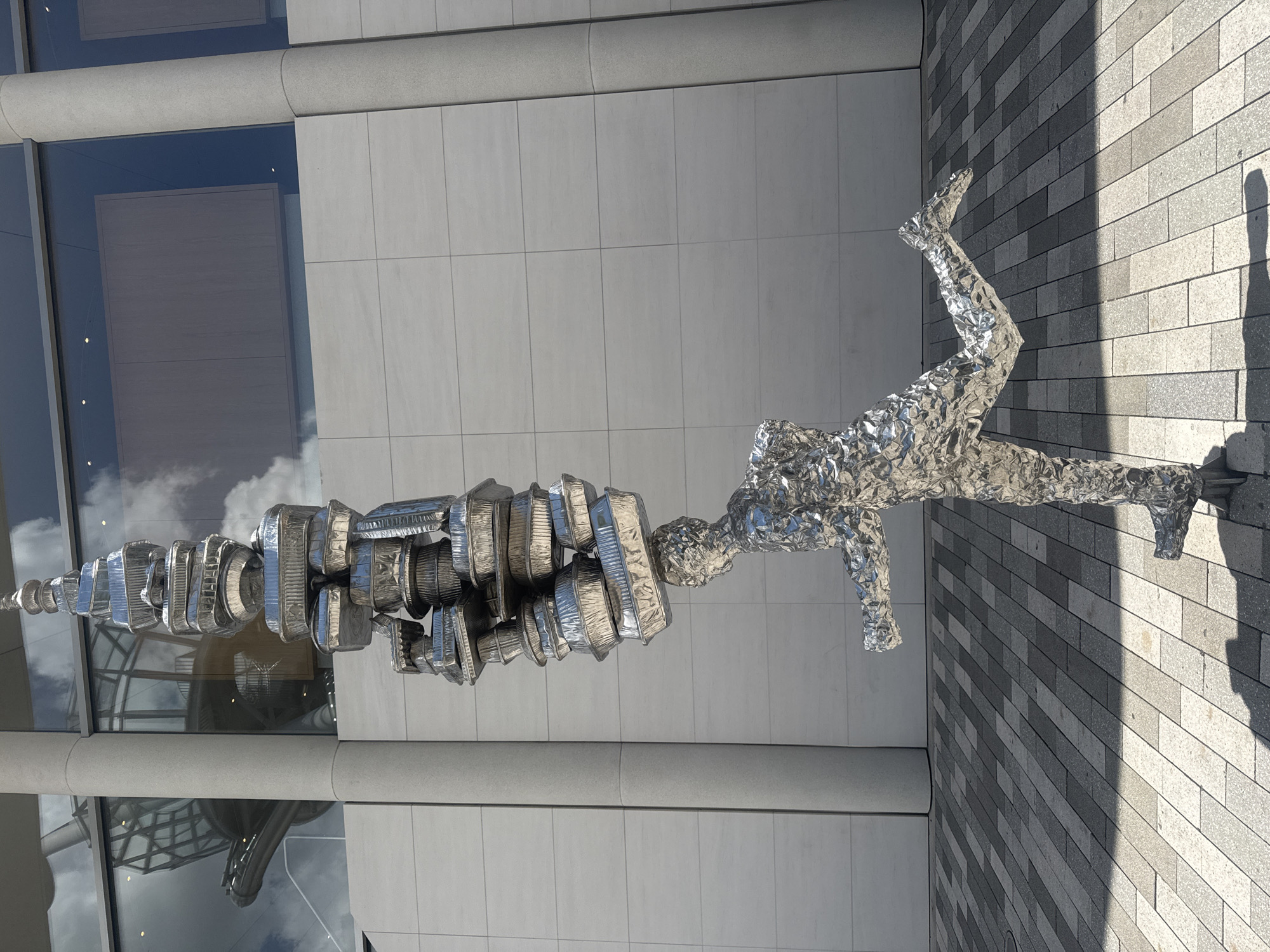 The image captures a public art installation located in Aventura, United States of America. The sculpture consists of a human figure made of reflective material (likely aluminum or stainless steel) posed in a dynamic, almost athletic position, seemingly running or leaping. Connected to the head of the figure is a large, abstract form also constructed of reflective material. This form is segmented, resembling a series of stacked or connected cylinders or disks, potentially alluding to industrial or mechanical components. The sculpture is situated outdoors, with a modern building featuring large glass windows and a grey tiled or paved ground in the background. The building's windows reflect the sky, suggesting a partly cloudy day. A shadow is cast on the ground, indicating a light source from the right side of the frame. The image appears to have been taken from a low angle, emphasizing the height and presence of the sculpture.