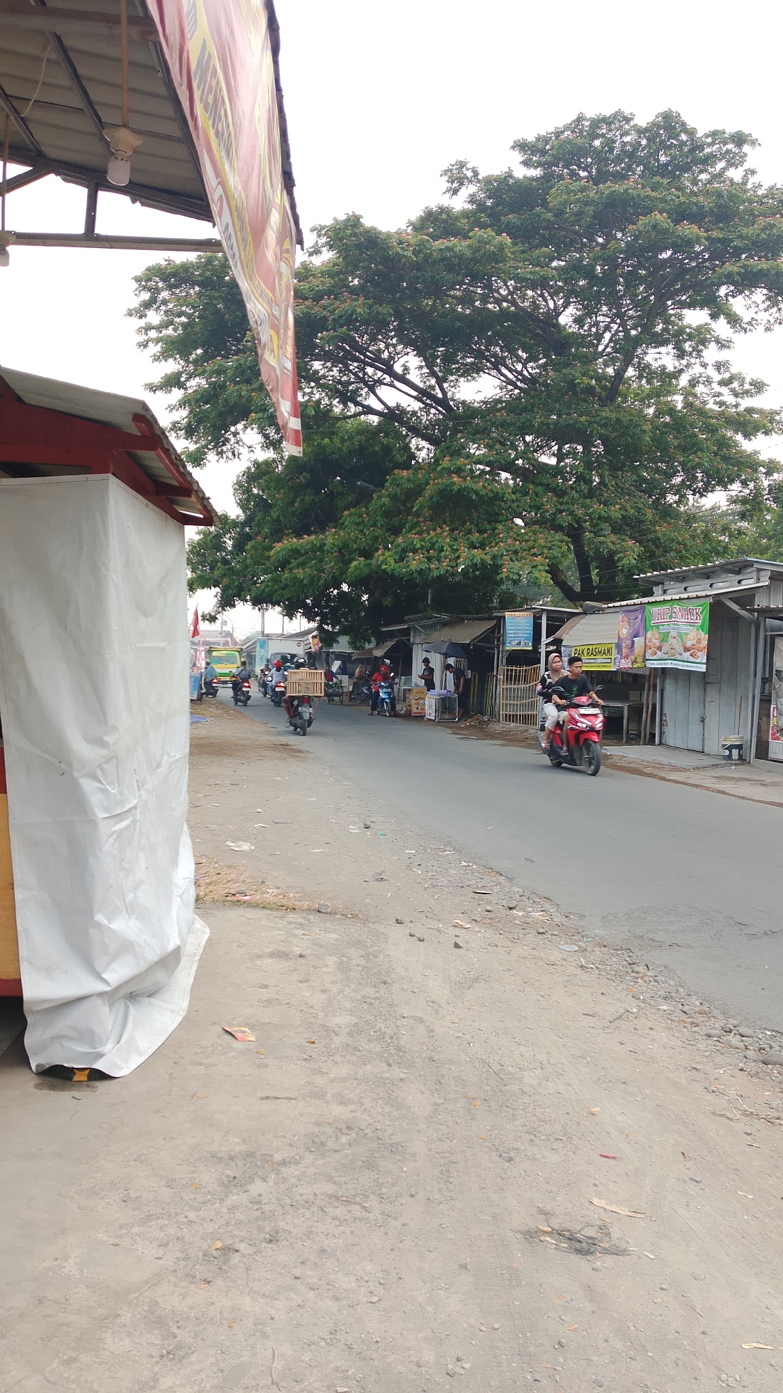 This is a street scene in Pangkah, Indonesia, featuring a road lined with small shops and large trees. On the left side of the image, a white tent or cover is visible in the foreground, part of a temporary structure. A banner hangs overhead on the left, partially obscuring the sky. The road itself has several motorcycles on it, with riders and passengers. One motorcycle, a red one in the middle of the road, carries two people. Another motorcycle further back has a wooden crate strapped to it. In the background, a yellow truck is visible, and more motorcycles and people are scattered among the shops. The shops appear to be small, open-air businesses, some with signage. The environment is somewhat dusty, with a dirt shoulder along the road. The overall impression is of a busy, local marketplace or roadside commercial area.