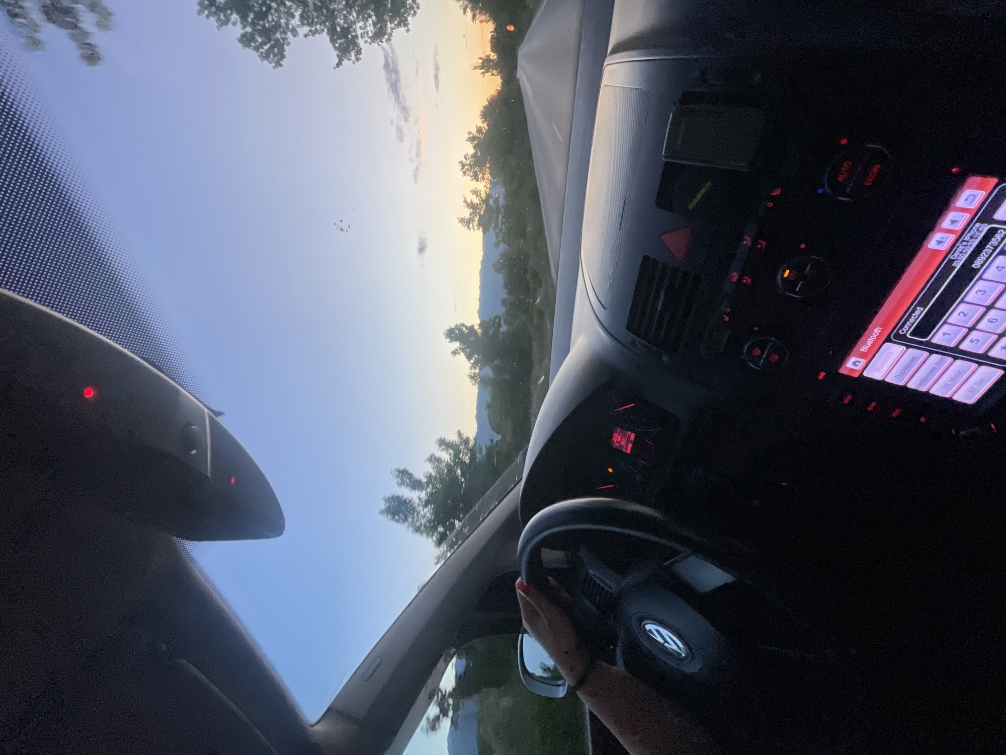 The image, taken inside a car, captures a person driving on a road in Dabnitsa, Bulgaria, as dusk approaches. The camera is positioned to show the dashboard and the driver's hand on the steering wheel, with the road ahead visible through the windshield. The sky displays the soft hues of sunset, with scattered clouds. Trees and distant mountains line the road. The car's interior features a digital display on the stereo system, showing "Bluetooth" and "Connected" status, along with a phone number and a contact name. The steering wheel has the Volkswagen logo. Two red indicator lights are visible on the overhead console.