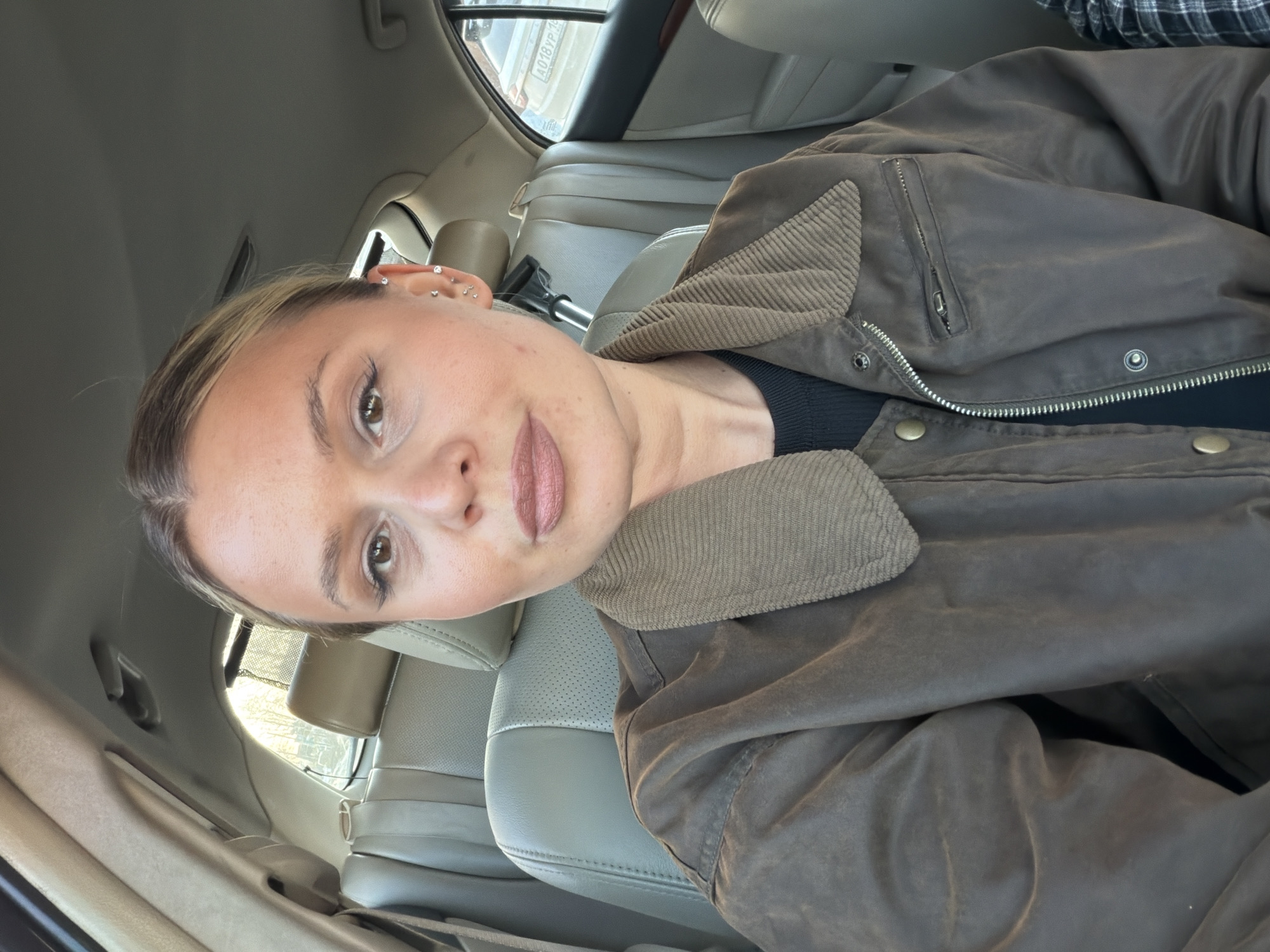 The image shows a woman in the driver's seat of a car. She has light brown hair pulled back, multiple piercings in her ear, and is wearing a brown jacket with a corduroy collar over a black shirt. The interior of the car is light gray. The car is in Sarapul, Russia, as the license plate of a car visible through the window is registered in Udmurtia.