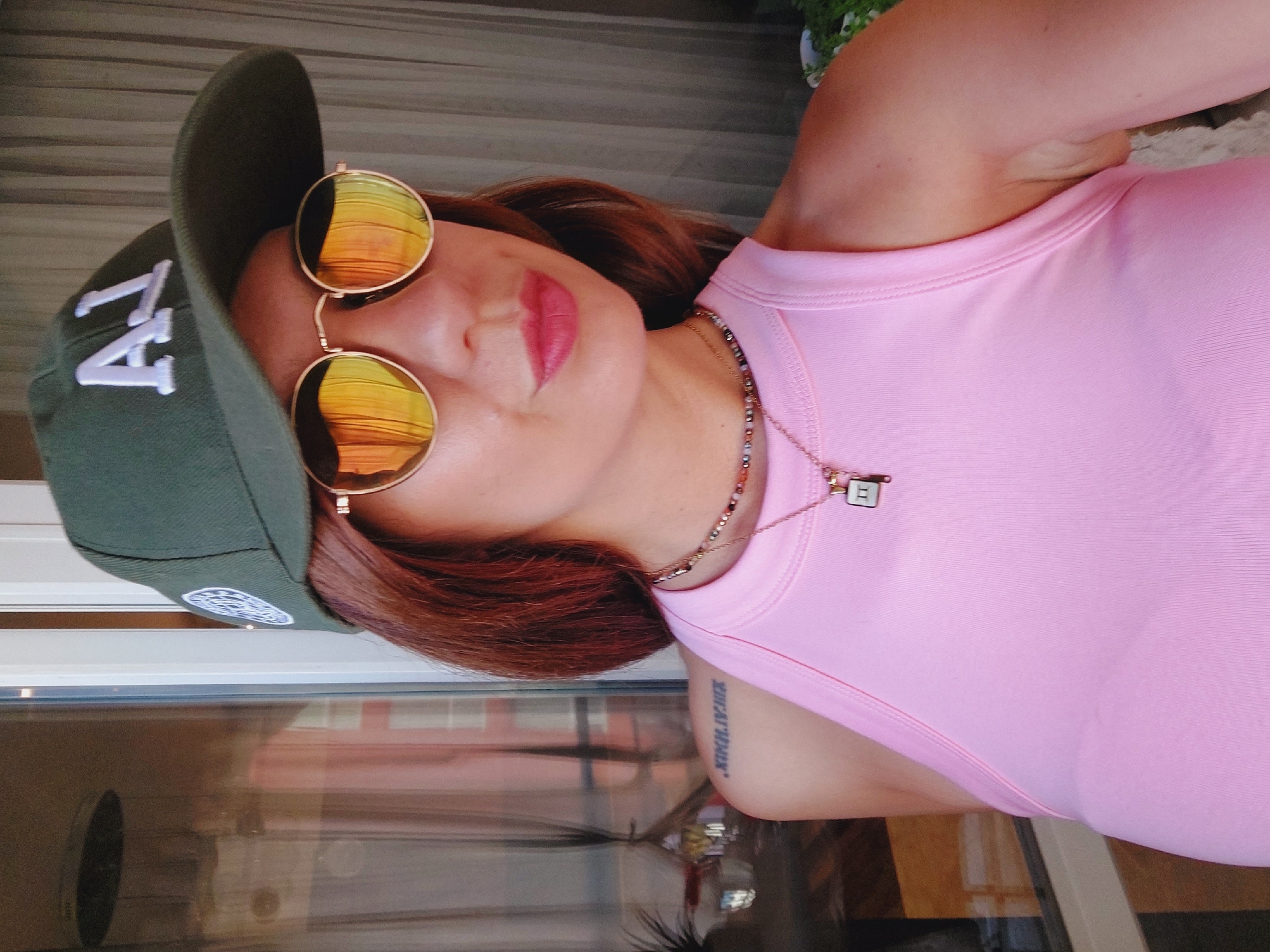 A woman with auburn hair, wearing a dark green baseball cap with white "LA" lettering, mirrored sunglasses with orange lenses, a beaded necklace with a small pendant, and a pink tank top stands in front of a window. Her left shoulder bears a tattoo of Roman numerals. The environment appears to be indoors, with a window reflecting an outdoor scene.