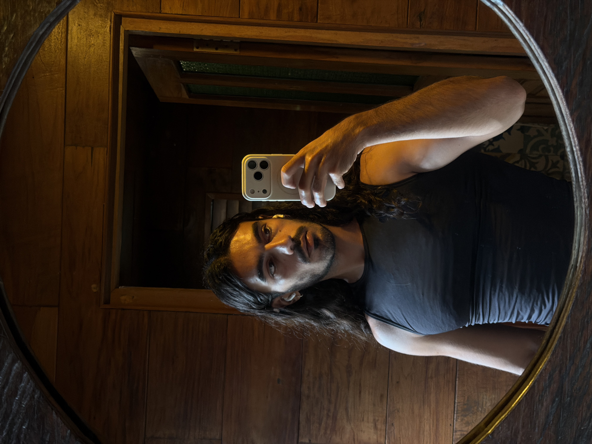 A man with long dark hair and a trimmed beard is taking a selfie in a round mirror. He is holding a white smartphone with a triple camera setup in his right hand. The man is wearing a black tank top. The reflection shows a wood-paneled wall behind him with a small window opening at the top. The scene appears to be indoors, with warm, ambient lighting. The image was taken in North Kuta, Indonesia.