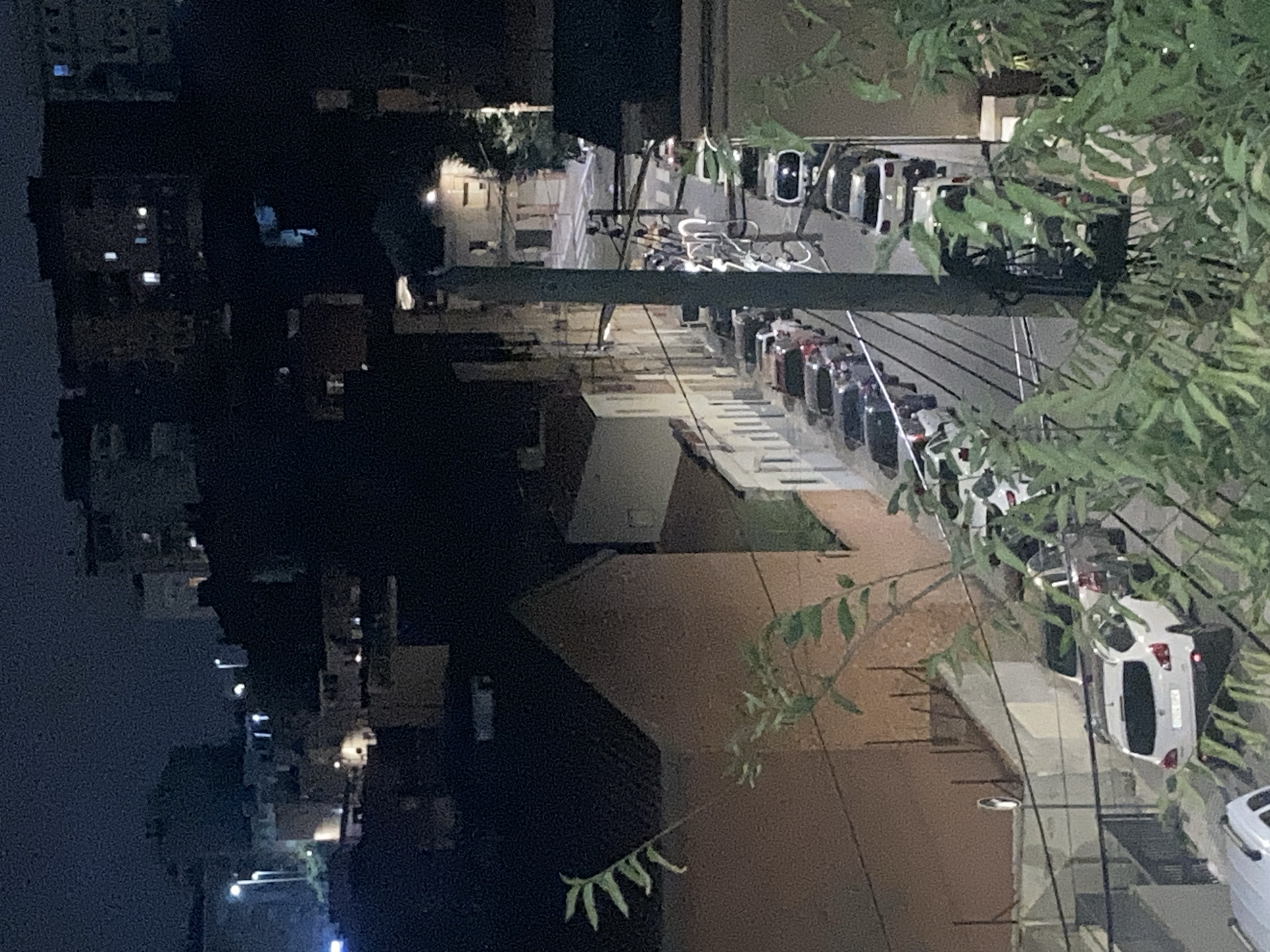 The image depicts a nighttime view of a narrow, sloped street in the city of Sabadell, Spain, likely in a residential neighborhood given the presence of apartment buildings. The street is lined with parked cars on both sides, creating a sense of density. A few streetlights are visible, casting a dim glow on the scene. The buildings themselves appear to be multi-story, with some windows illuminated, suggesting that people are inside. There are no visible individuals actively engaged in any discernible actions, but the overall impression is of a quiet, urban evening. The foreground is partially obscured by foliage, hinting at a vantage point from a higher elevation or a nearby garden.