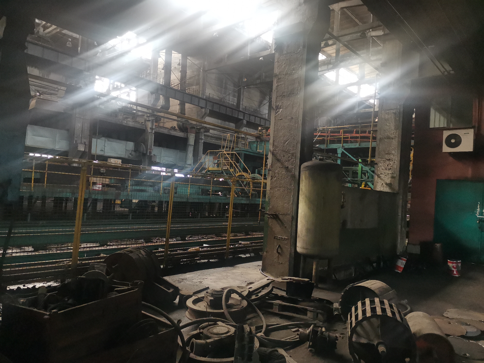 The image depicts the interior of an industrial facility, possibly a factory or mill. Light streams in through high windows, creating strong light and shadow patterns. The foreground contains a collection of discarded machine parts, including wheels, gears, and other components, strewn on the floor. Some of these parts are contained within a wooden crate. In the middle ground, there's a green and yellow metal structure with a fence and a staircase, suggesting an elevated walkway or platform. To the right of this structure stands a large, cylindrical tank or silo. A closed, green door is visible further right, with what appears to be an air conditioning unit above it. The background shows more of the industrial structure, with beams, pipes, and machinery. The overall atmosphere is one of disuse, decay, and neglect. There is no verifiable information to confirm the location within Zhivkovo, Bulgaria. The scene does not show any people.