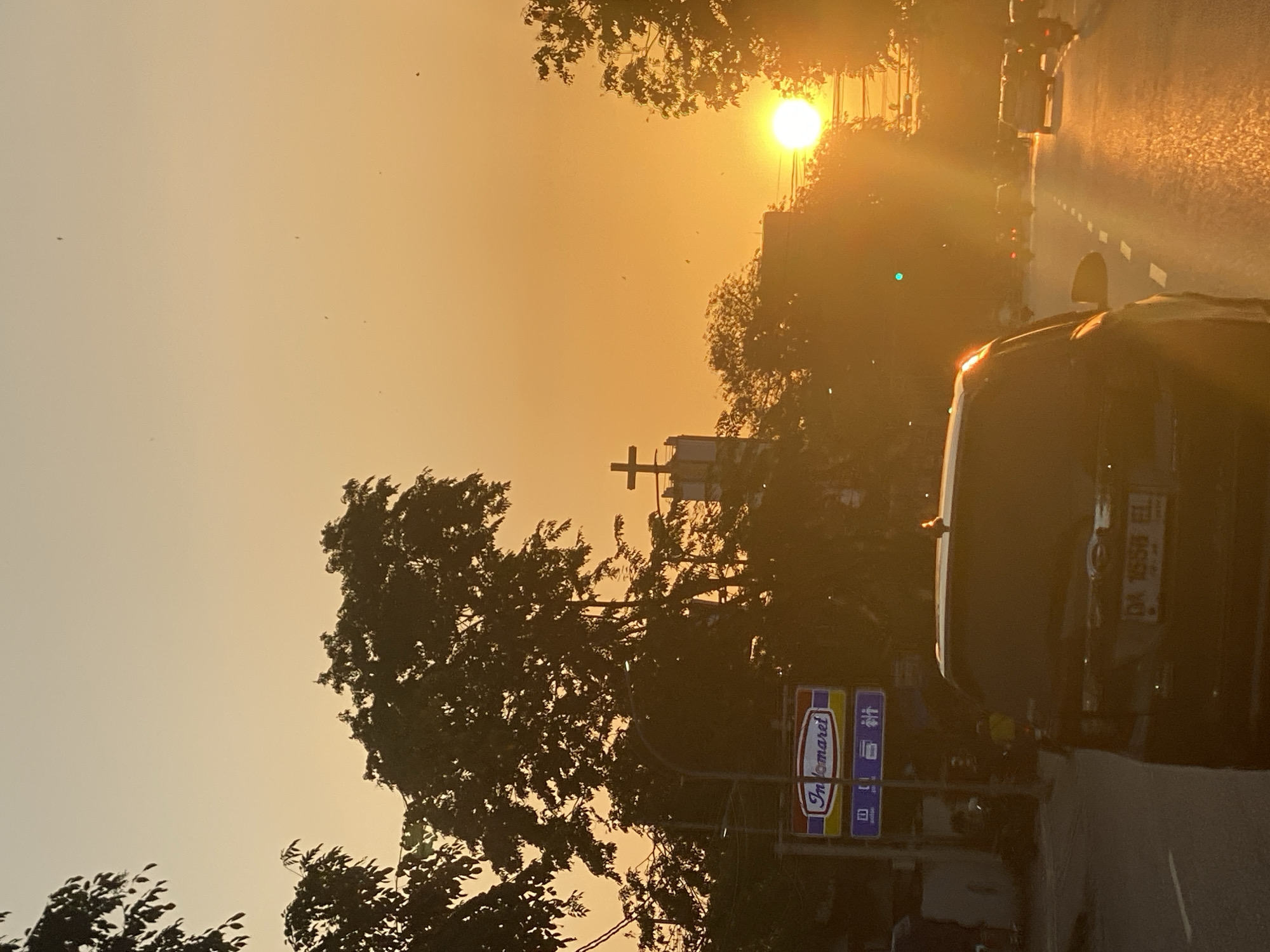 This is a photograph of a street scene in Banjarmasin, Indonesia, during sunset. The sky is a gradient of warm orange and yellow hues, with the bright sun low on the horizon, casting a warm glow over the scene. Silhouetted trees frame the shot, some appearing dense and leafy, others more sparse. A prominent feature is a building topped with a cross, suggesting a religious institution, possibly a church.
In the foreground, the rear of a dark-colored SUV is visible, its license plate reading "DA 1656 EL." The SUV is on a paved road, which reflects the golden light of the setting sun. Further down the road, other vehicles, including motorcycles and cars, are also present, suggesting moderate traffic. The road is marked with a white dashed line. To the left of the road, a sign for "Indomaret," a convenience store chain common in Indonesia, is clearly visible, indicating the urban setting. The overall atmosphere is tranquil, capturing the end of the day in a bustling Indonesian city.