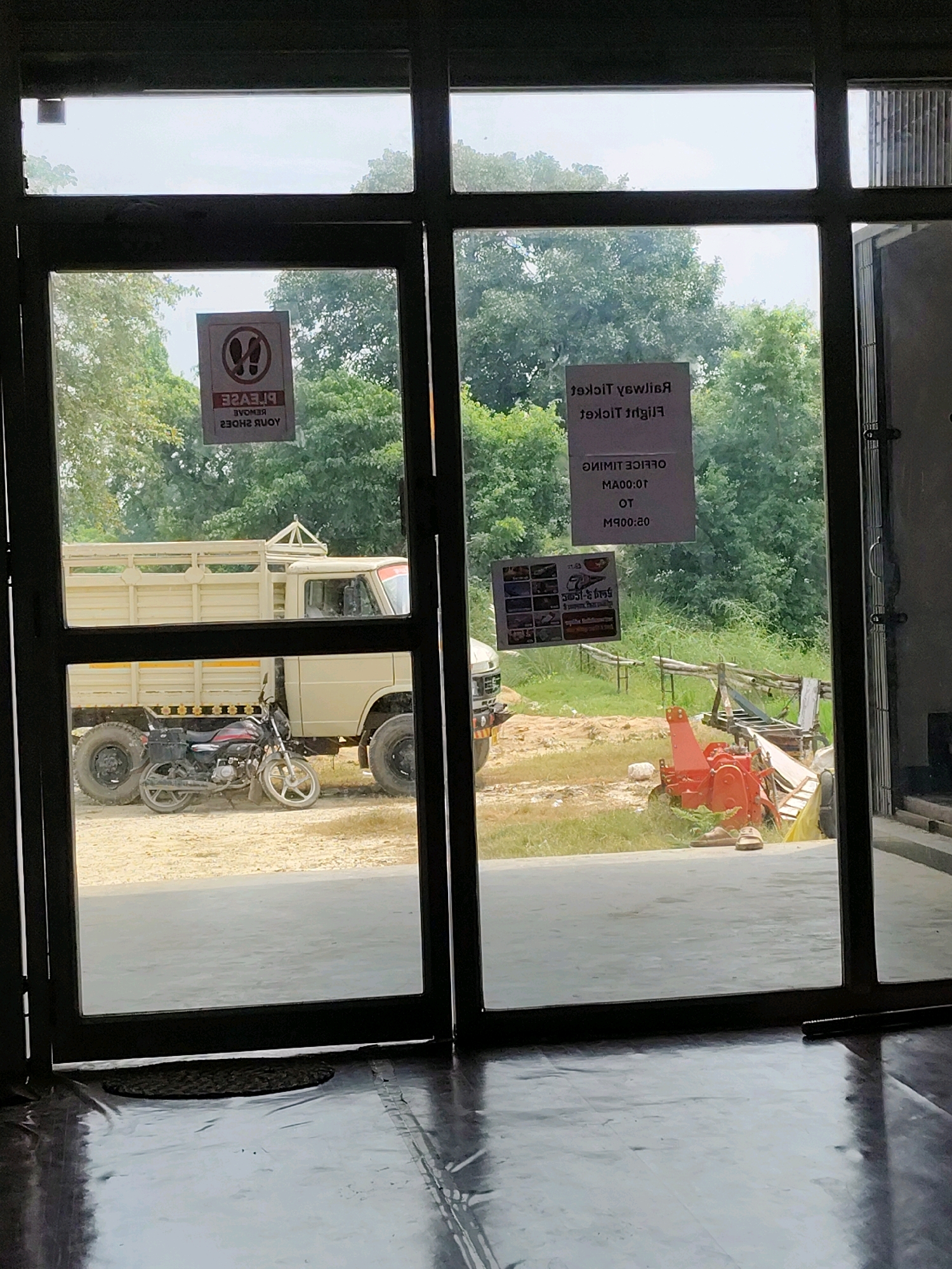 The image shows a view through a glass door of a scene in Harpur, India. Outside, a beige truck is parked next to a motorcycle. In the background, there are trees and a red piece of farm equipment. Two signs are visible on the glass door; one requests that shoes be removed, and the other displays office hours for railway and flight tickets. The floor inside the building is dark and reflective.