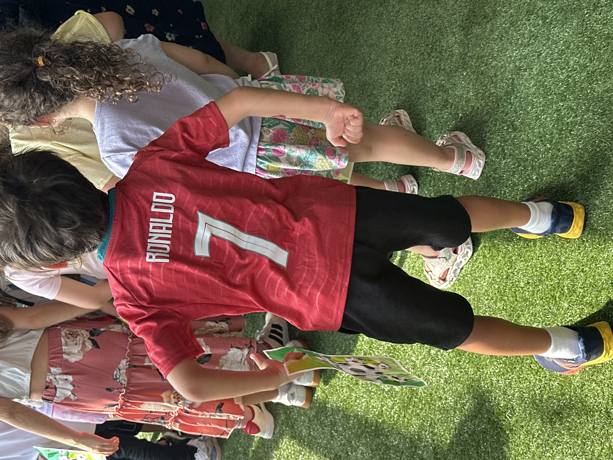The image shows the backs of several children standing on a green, artificial turf surface. The child in the foreground wears a red t-shirt with "RONALDO" and the number "7" on the back. This child is also wearing black shorts, white socks, and blue and yellow sneakers. To the right of this child is another child wearing a light purple t-shirt and a colorful skirt with a fruit and floral pattern. Further to the left, partially visible, is a child wearing a white top and a long, pink skirt with a floral pattern. Another child, in the far background, wears a dark blue dress with a floral pattern. The scene appears to be an outdoor event, possibly a party or gathering, and the children are engaged in an activity as evidenced by one child holding a colorful paper item. The artificial turf suggests an urban setting, possibly a park or recreational area. No specific New York City landmarks are visible.
