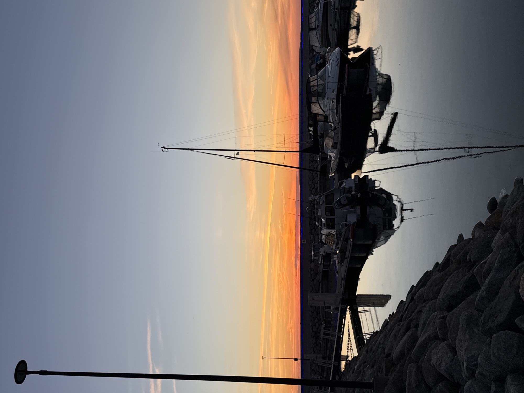The image depicts a marina at dusk, likely in Tallinn, Estonia, given the aesthetic. The sky is a gradient of pale blue at the top, transitioning to a vibrant orange and yellow sunset near the horizon. Wispy clouds are visible in the upper portion of the sky.
In the foreground, to the left, is a dark silhouette of a tall lamppost. Below the lamppost, a rocky embankment runs along the edge of the water. A wooden pier extends into the water, with several boats moored to it. These boats are silhouetted against the brighter sky, and their reflections are visible on the calm water.
One prominent sailboat with a tall mast stands out, its rigging also reflected in the water. Other motorboats with enclosed cabins are also docked. The water is dark and still, creating clear reflections of the boats and the pier. A faint blue line is visible across the horizon, possibly indicating distant lights or structures. The overall atmosphere is serene and peaceful, characteristic of a quiet evening in a harbor. There are no people visible in the image.