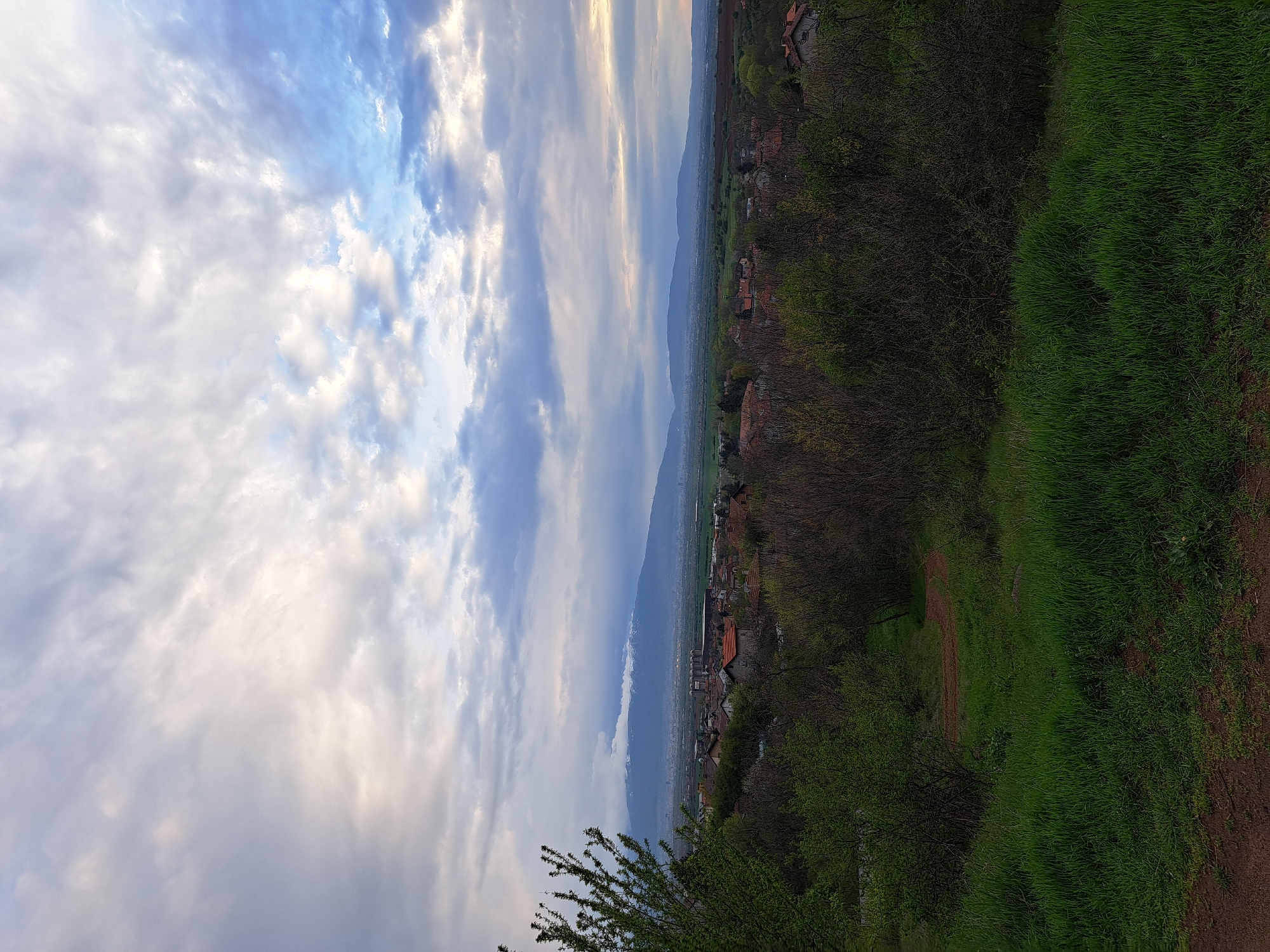 The image captures a serene landscape from an elevated viewpoint in Lokorsko, Bulgaria, showcasing a rural village against a backdrop of mountains and a dynamic sky.
In the foreground, on the right, a vibrant expanse of fresh green grass is visible, suggesting spring or early summer. A faint dirt path winds through this grassy area. On the left, dense trees and bushes, some with emerging green leaves and others still appearing somewhat bare, frame the immediate area, casting some shadows.
The midground features a charming village or small town spread across the valley. Numerous houses with distinct reddish-brown tiled roofs are interspersed among trees and greenery, indicating a peaceful settlement. Beyond the village, the land stretches out towards a distant horizon.
In the background, a formidable mountain range dominates the vista. The mountains appear somewhat hazy or partly obscured by atmospheric conditions, possibly low-lying clouds or mist, contributing to a sense of depth and scale.
The sky is a prominent element, filled with a diverse array of clouds. Patches of clear blue sky are interspersed with fluffy white cumulus clouds and broader, softer grey and white stratocumulus formations. Some clouds are touched with warm, golden light, suggesting either late afternoon or early morning, with the overall lighting leaning towards an impending sunset.
There are no people, specific activities, or visible text. The scene evokes a sense of calm and the beauty of a natural and inhabited landscape.