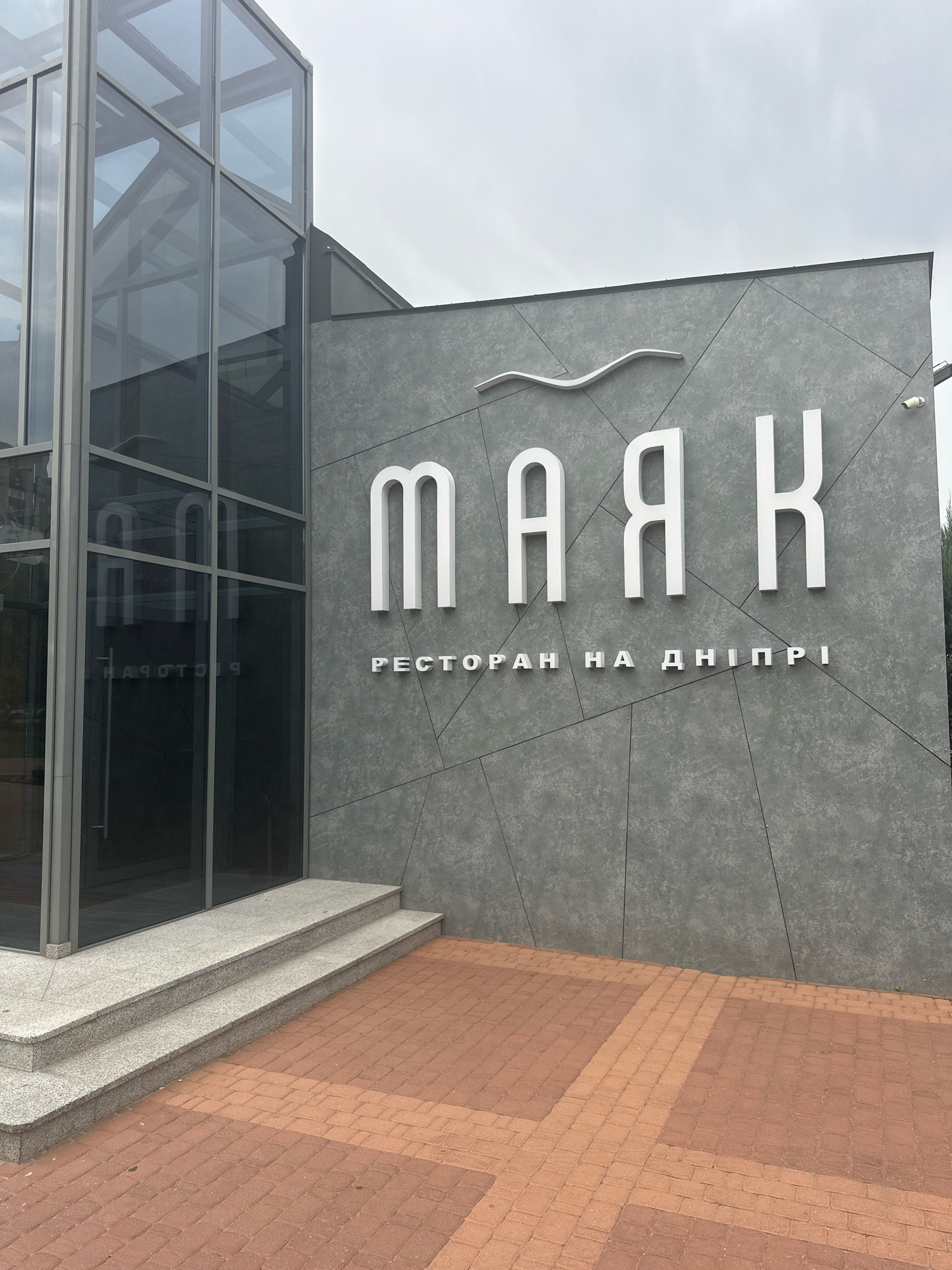 An outdoor shot captures the facade of a building with a modern design. A large glass entryway with gray metal framing is on the left, with reflective glass showing a reversed sign. Two gray granite steps lead up to the entrance. To the right of the entryway, the building's exterior is clad in gray panels with a textured finish, arranged in a geometric pattern. Prominently displayed on the gray facade are large, white, three-dimensional letters spelling "МАЯК" in a clean, sans-serif font. Below this, smaller white letters spell "РЕСТОРАН НА ДНІПРІ". A subtle wavy white design element is placed above the large letters. A white security camera is mounted on the upper right side of the building. The ground in front of the building is paved with reddish-brown interlocking bricks. The sky above is overcast.