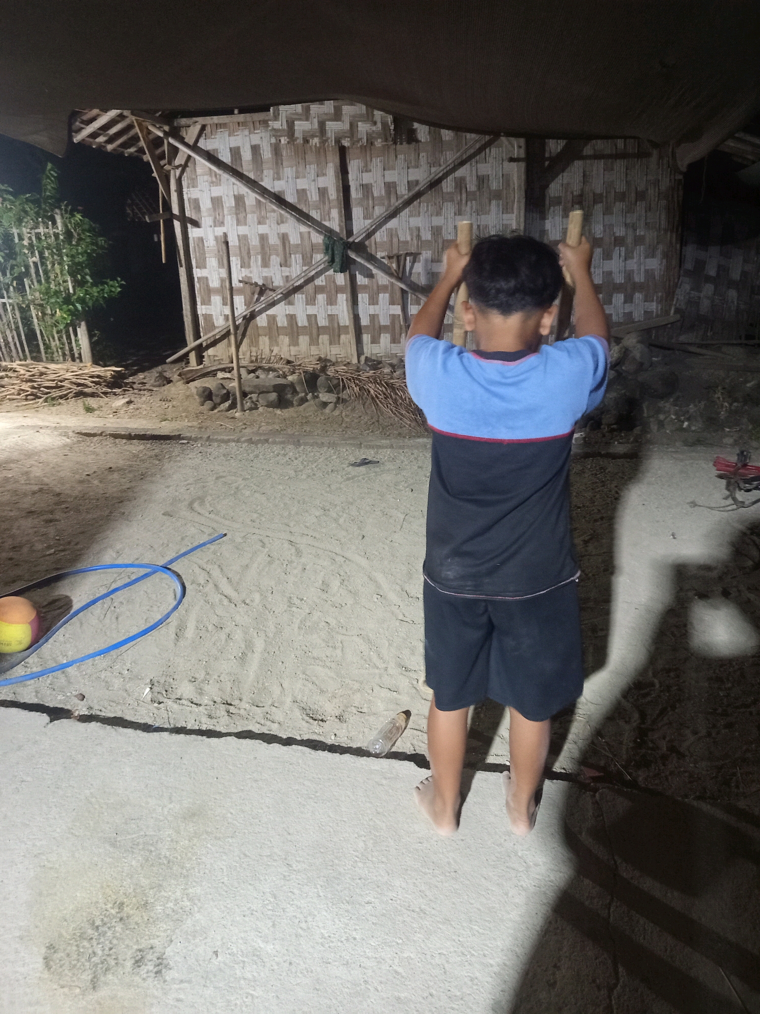 A young boy is captured from behind, mid-action, while seemingly hanging from two wooden poles. He is wearing a blue and black t-shirt with red trim and black shorts. His feet are bare and dangling in the air. The scene appears to be outdoors, possibly in the city of Kendel, Indonesia. Behind him, a rustic structure made of woven bamboo and wood is visible, with a thatched roof. The ground is a mix of sand and concrete. A blue hose and a partially visible ball are on the sand to the left of the boy. The lighting suggests it is either dusk or night, with shadows cast by the boy and other unseen objects. The overall environment suggests a rural or semi-rural setting.