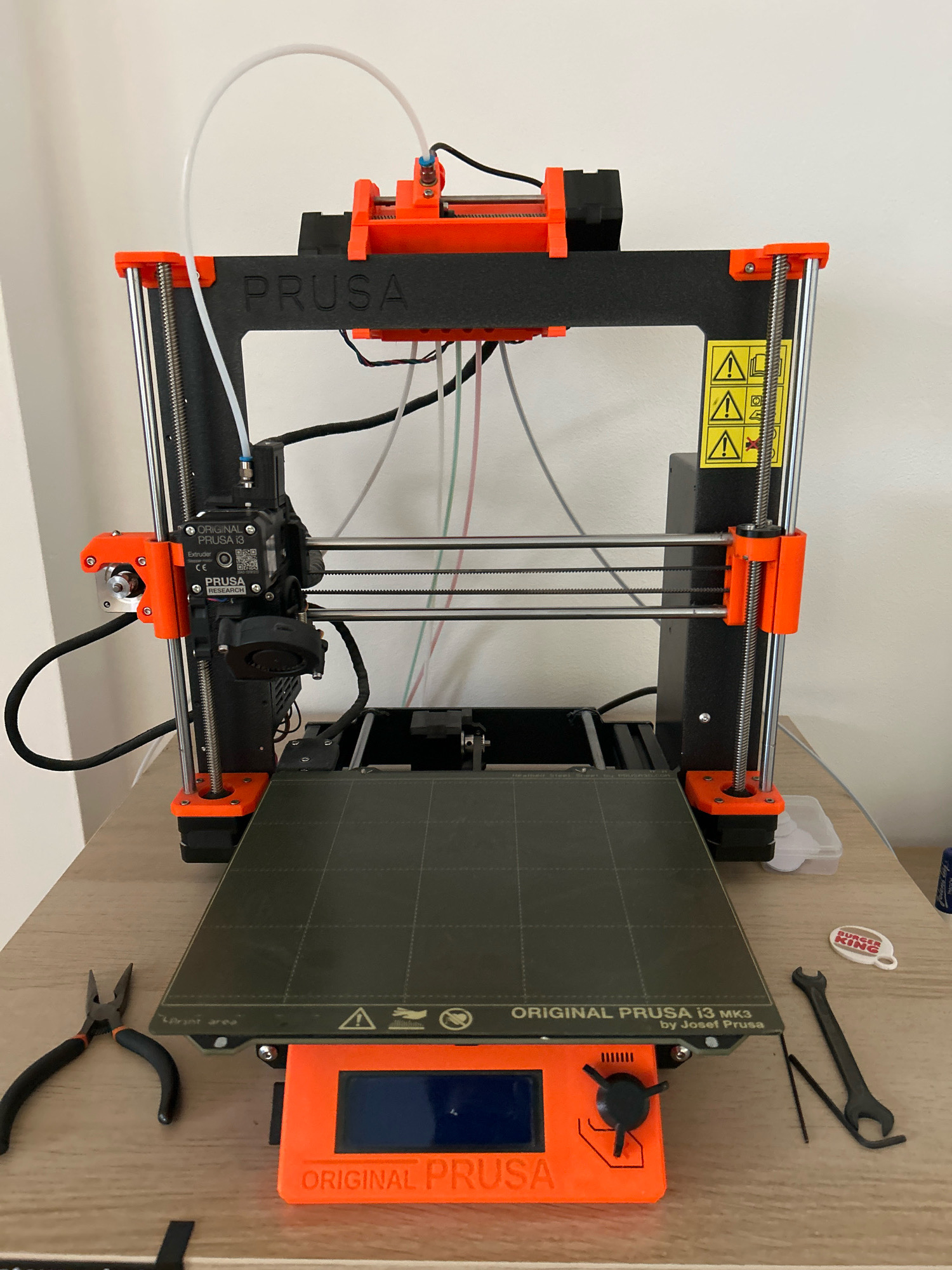 The image shows a Prusa i3 MK3 3D printer placed on a wooden desk in what appears to be an indoor setting. The printer is black with orange accents and has its build plate, extruder, and various wires visible. To the left of the printer, a pair of black-handled pliers are positioned on the desk. To the right, a black wrench, a small Allen key, and a Burger King keychain are present. The background consists of a plain white wall. The scene is well-lit, suggesting it was taken in natural or artificial light. No people are visible in the image. The location is stated to be Ořechov, Czechia.