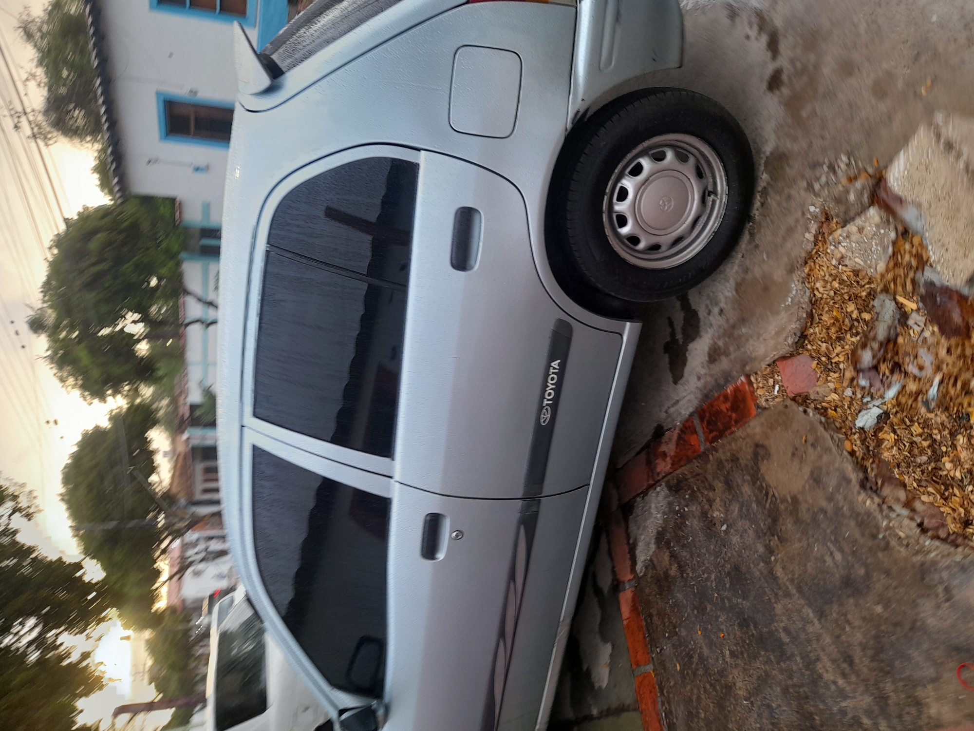 The image shows a silver Toyota car parked on a wet, cracked concrete surface bordered by red bricks. The car has tinted windows and a small spoiler on the rear hatch. A white car is partially visible in the background, along with some trees and houses. The sky is overcast, suggesting it may have rained recently. The scene appears to be a residential street in Santa Ana, Venezuela.