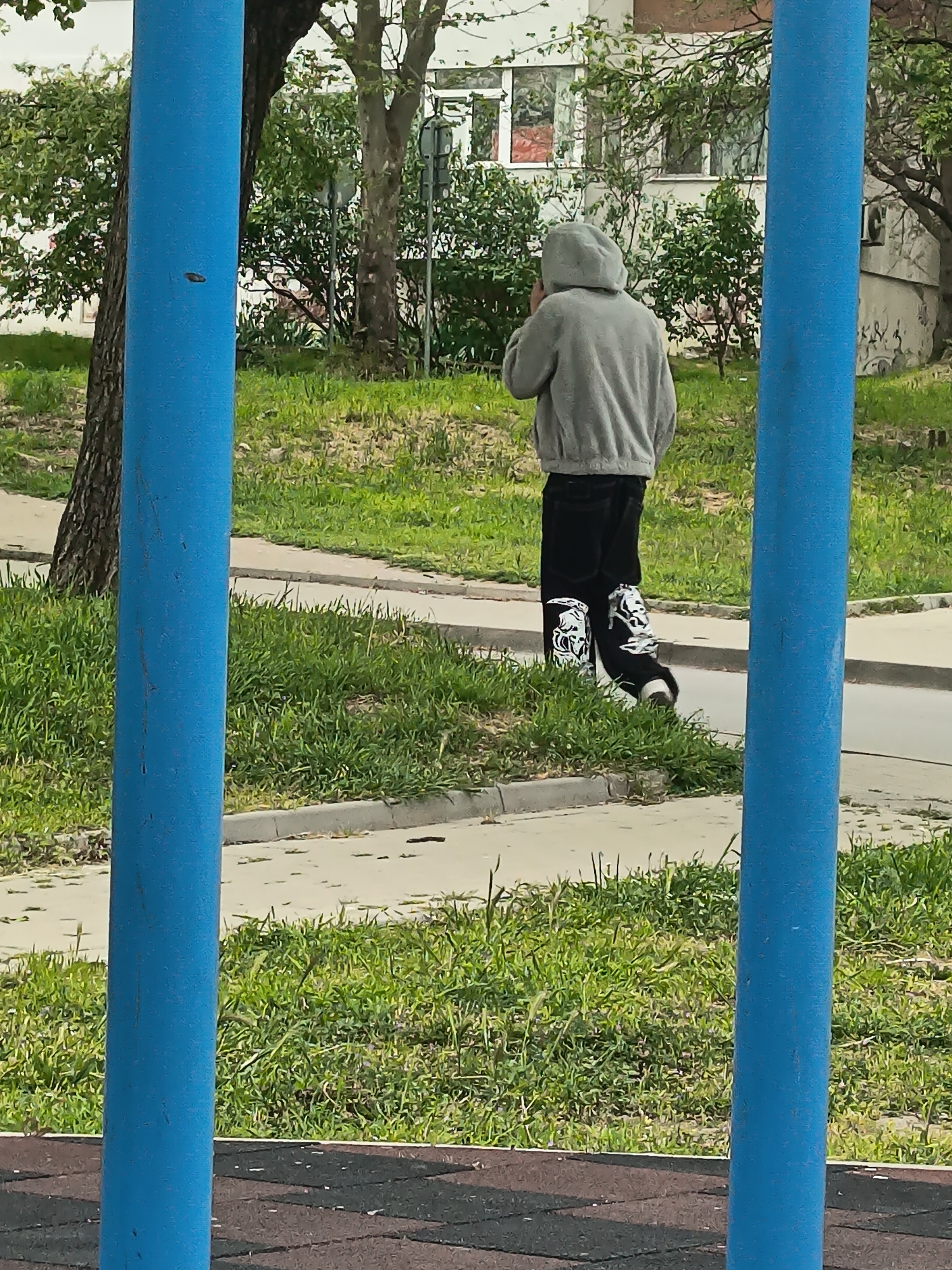 A person wearing a grey hoodie and black pants with white designs is walking away from the camera on a sidewalk adjacent to a grassy area in Varna, Bulgaria. The person is positioned between two blue metal poles. A tree is to the left, and a building is in the background.