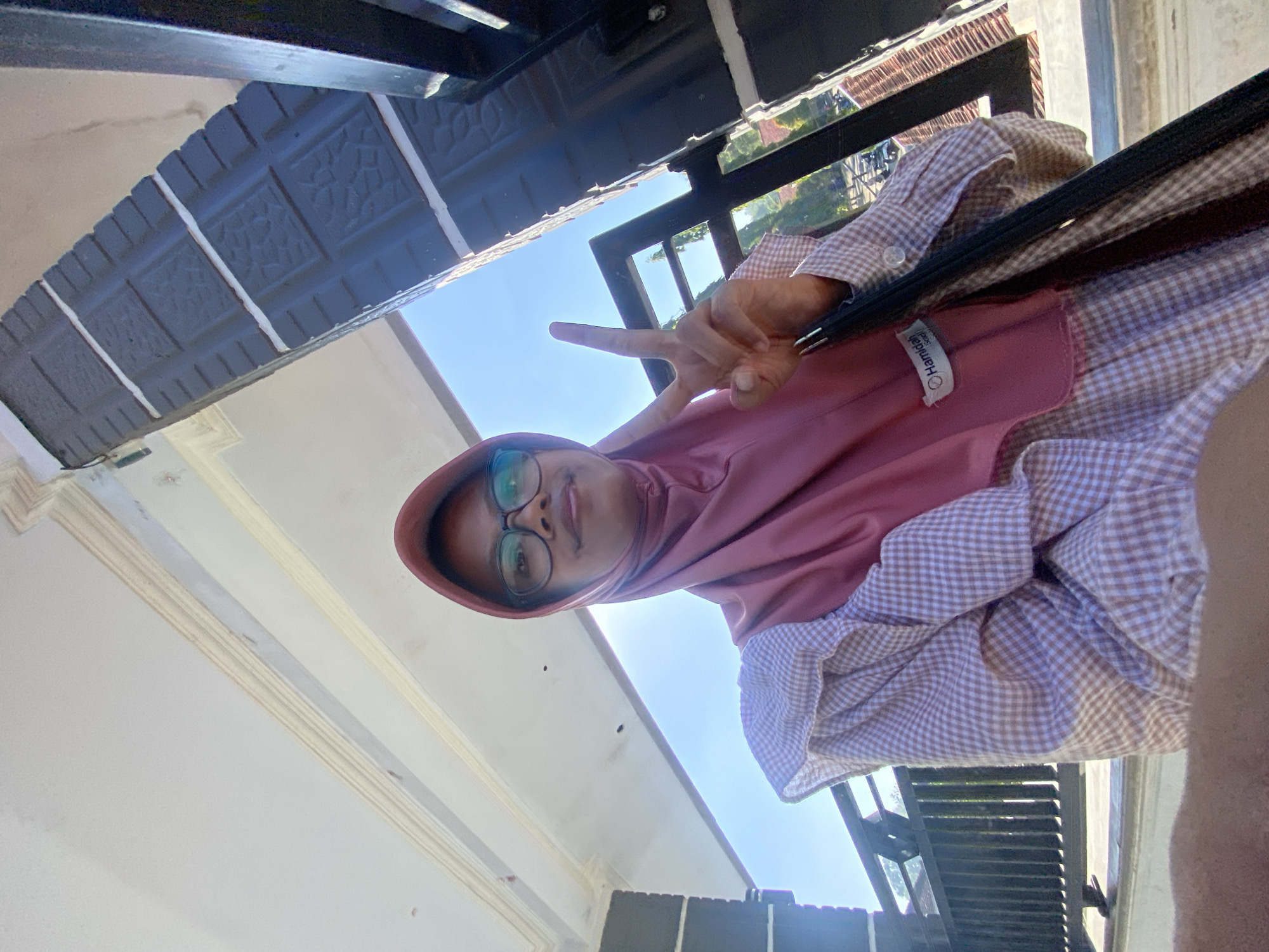 A young woman wearing a pink hijab and glasses is taking a selfie. She is wearing a light brown and white checkered shirt with puffed sleeves. She is holding up two fingers in a peace sign. Behind her, there is a white ceiling with decorative molding, a dark gray pillar with textured square patterns, and a black metal railing. The sky is blue and clear, suggesting it is daytime. The scene appears to be outdoors, possibly on a porch or balcony of a building in the city of Gunungsari, Indonesia.