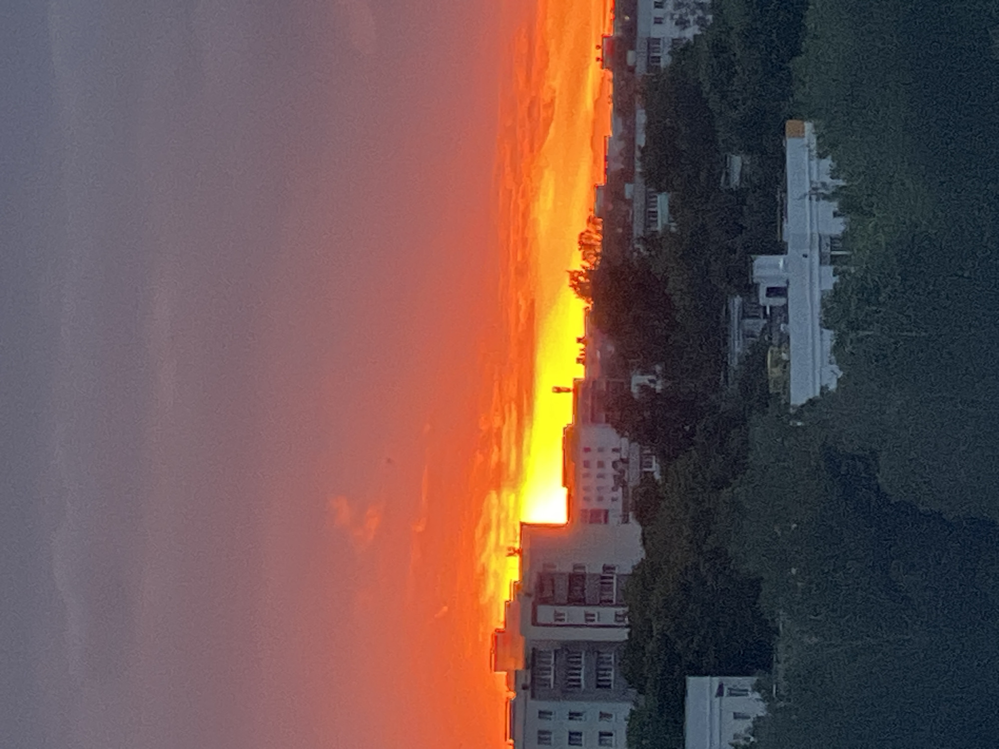 A vibrant sunset casts an orange and red glow over a city skyline. In the foreground, a dense layer of dark green trees obscures the lower portions of buildings. Above the trees, several multi-story apartment buildings are visible, some with distinctive architectural features such as balconies and window patterns. The sky transitions from a deep purple at the top to intense orange and yellow hues near the horizon, where the sun is setting, partially obscured by clouds and buildings. The scene depicts an urban environment at dusk, characterized by the dramatic light of the setting sun illuminating the sky and silhouetting the city's architecture. The specific location in the city of Minsk, Belarus, is not determinable from the provided image alone.