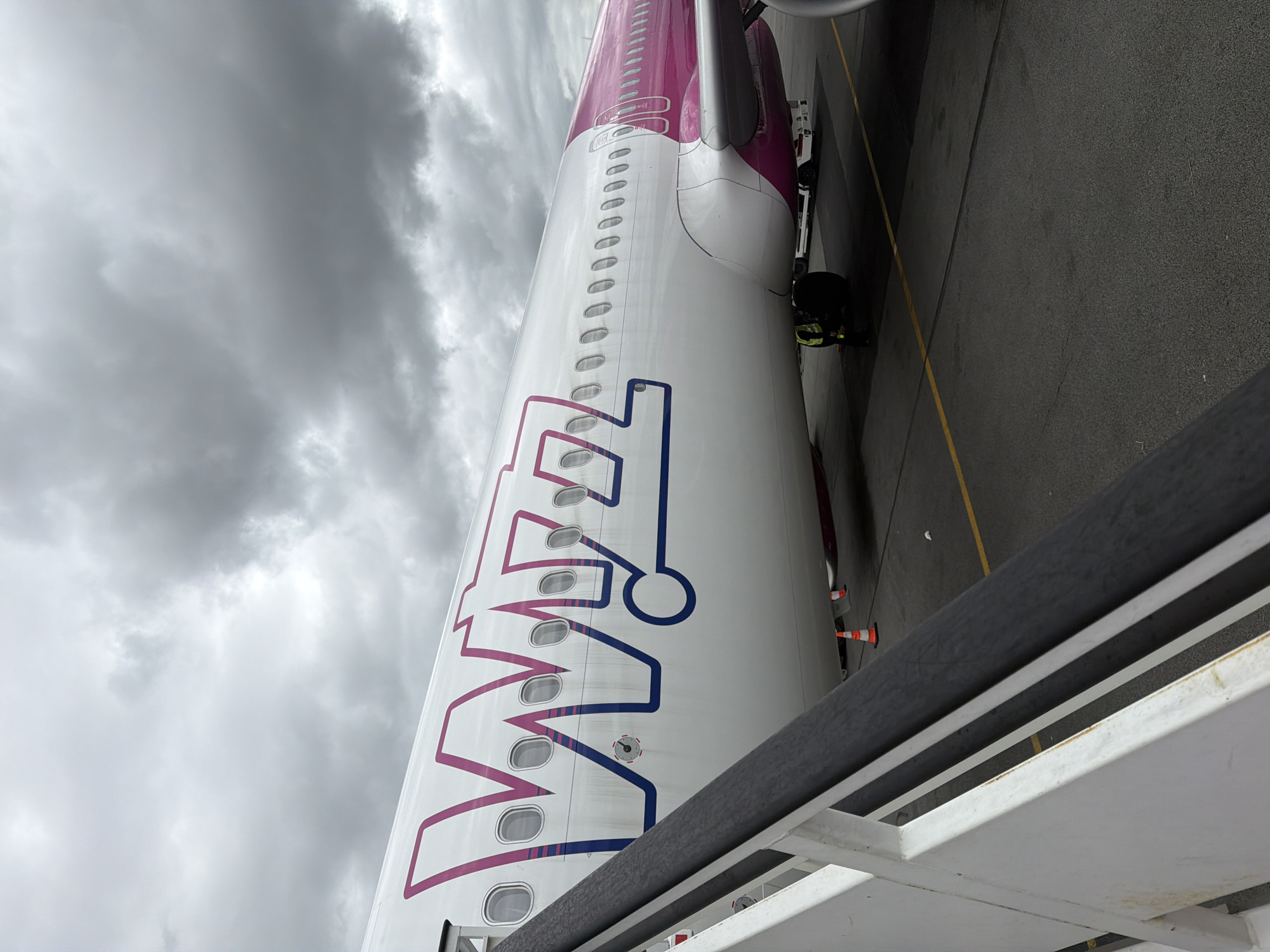 A large passenger airplane, specifically a Wizz Air aircraft, is prominently featured, parked on an airport tarmac under a heavily overcast sky. The airplane's white fuselage, adorned with the distinctive pink and blue "WIZZ air" logo, occupies the central and left portions of the frame. Numerous oval windows are visible along the side of the aircraft.
In the foreground, a white and dark grey railing, likely from a mobile staircase or passenger boarding bridge, cuts diagonally across the bottom right. On the tarmac, a yellow line marks a boundary, and a person wearing high-visibility clothing is visible near the plane's underside, suggesting ground operations are in progress. Further back, part of an engine or wing and ground equipment, including what appears to be the front of a service vehicle and orange safety cones, are seen near the aircraft's rear.
The sky is dominated by dark, heavy clouds, indicating an overcast or potentially stormy day. The scene depicts an airport apron, with the aircraft seemingly awaiting departure or having recently arrived at an airport in Tillé, France.