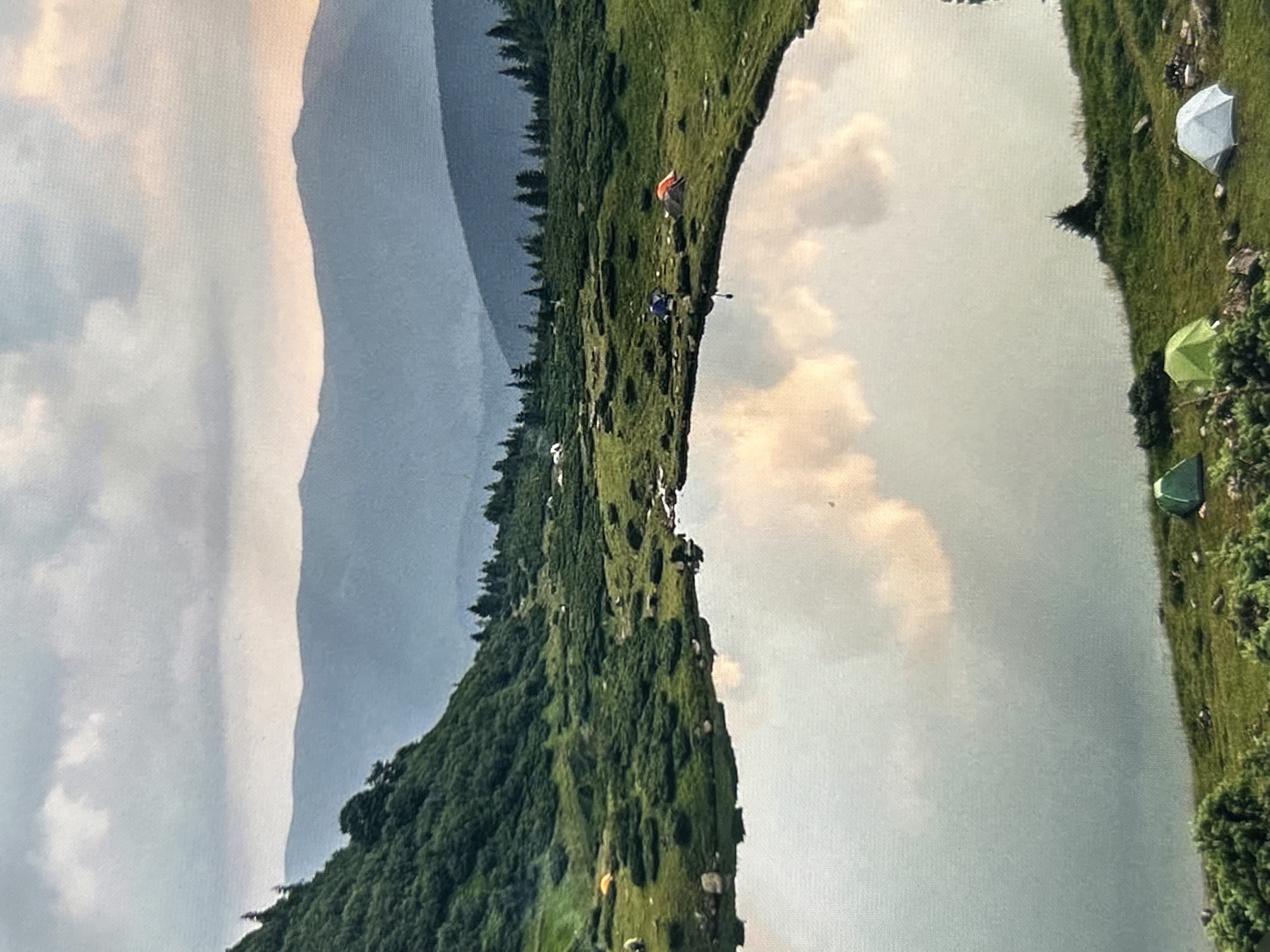 This is a scenic, eye-level outdoor photograph taken at dusk. The image depicts a tranquil alpine lake nestled within a lush, green mountainous landscape. The sky is overcast with dramatic clouds, some tinged with warm hues of orange and yellow from the setting sun, reflecting softly on the still surface of the lake.
The foreground is dominated by the reflective water of the lake, bordered by a grassy embankment. Several small camping tents are pitched along the shore, varying in color from green and lime to orange and white. Scattered rocks and low-lying green shrubs dot the landscape around the tents.
In the mid-ground, the terrain rises steeply to form a verdant, forested mountainside on the left, covered in dense green trees and shrubs. Further in the distance, rolling hills and mountains recede in layers, rendered in soft blue and grey tones due to atmospheric perspective. A thin line of coniferous trees marks the transition between the mid-ground and the distant mountain ranges.
The scene conveys a sense of peace and remoteness, characteristic of a wilderness camping spot. There are no people visible in the image. The photograph appears to be taken with a slight upward tilt, capturing the expanse of the sky and the layers of mountains. The location is not in the city of Uzhhorod, Ukraine, but rather a natural mountain environment.