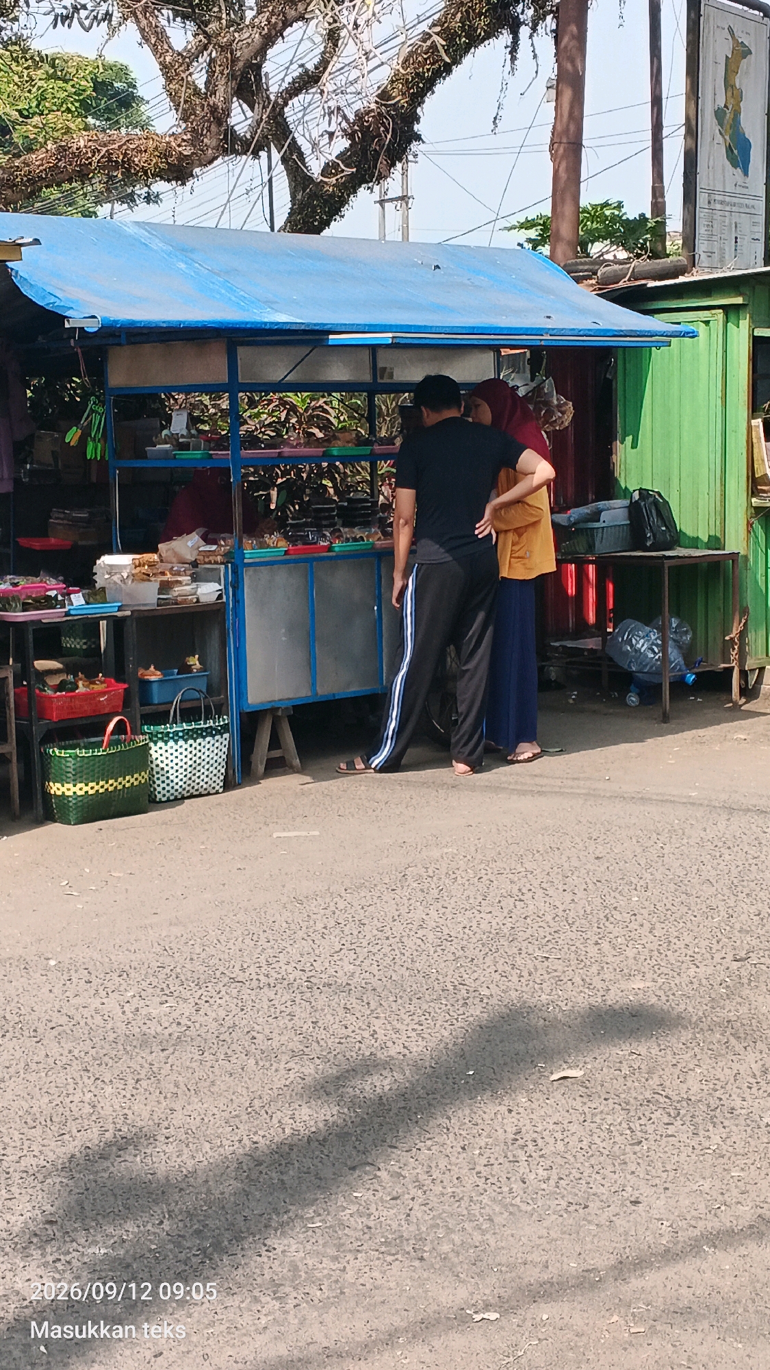 In the city of Tumpang, Indonesia, a street-side food stall with a blue tarpaulin roof is depicted. Two individuals, a man and a woman, are standing at the stall. The man, wearing a black t-shirt and black pants with white stripes, faces away from the camera. The woman, wearing a red hijab and an orange shirt, stands beside him, also facing away from the camera. They appear to be interacting with the stall, which displays various food items. The stall is positioned on a paved road with a tree overhead and a green building to the right. Several woven baskets are visible in front of the stall. The scene is captured during daylight, as indicated by the shadows on the road.