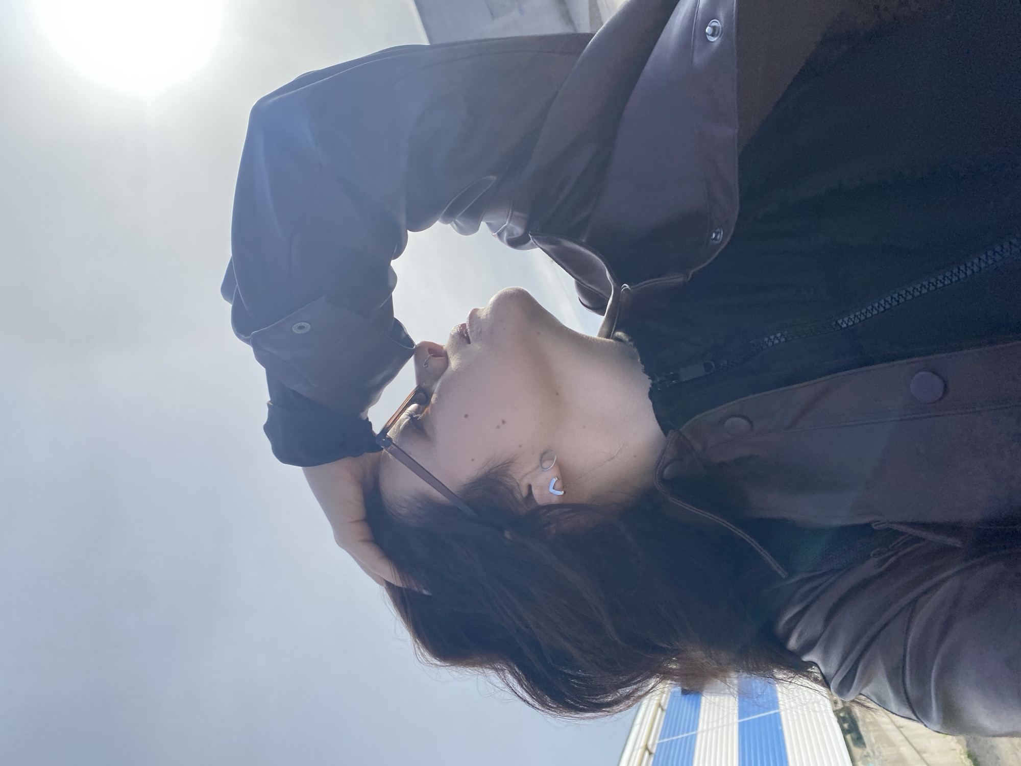 A woman with dark hair is viewed from a low angle, seemingly looking up at the sun. She wears glasses and earrings, and a dark jacket with a zipper and buttons. Her hand is near her head. The background includes a bright sky, possibly with the sun visible, and a glimpse of a building with blue and white stripes at the very bottom. It is not possible to confirm the location is Chaikovsky, Russia based on the visible content.