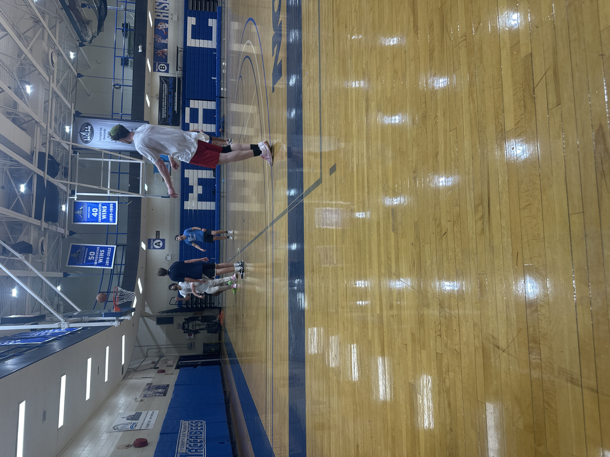 The image depicts a basketball court located in a gymnasium. Several individuals are present, some of whom appear to be engaged in basketball-related activities. Banners and signage on the walls indicate the court belongs to Yeshiva University, placing the location in New York City, United States of America. The court has the Yeshiva University Maccabees logo. The banners indicate winning seasons for men's basketball and men's tennis.
