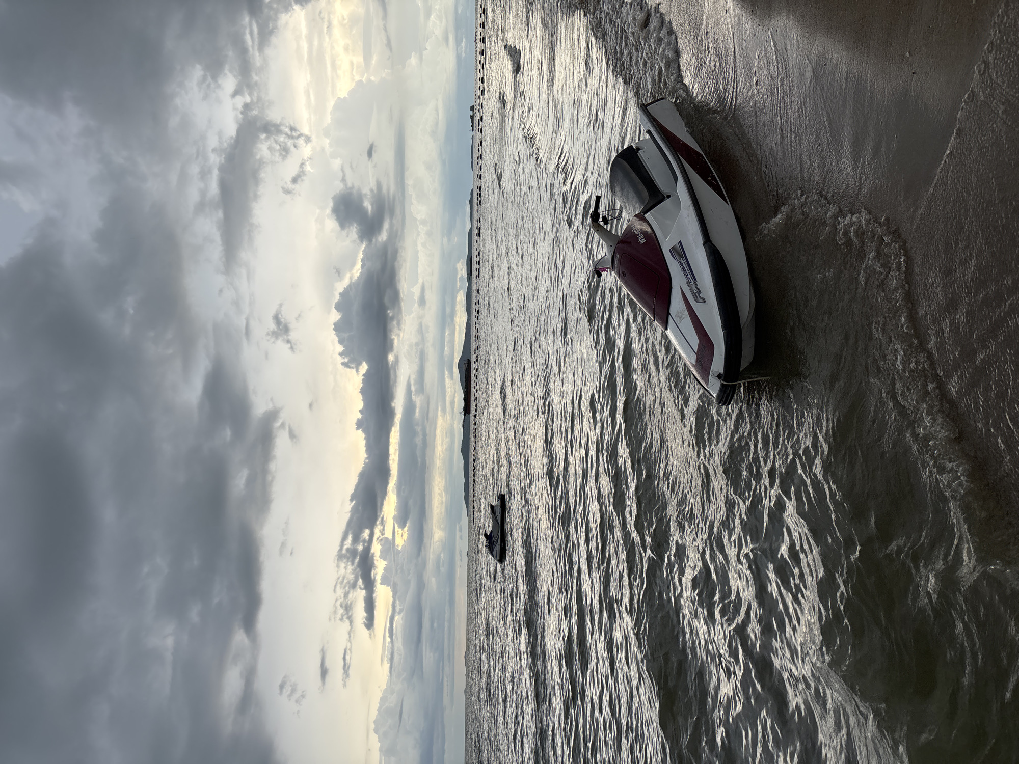 The image shows a jet ski beached on the shore of Pattaya City, Thailand, with another jet ski in the distance on the choppy water. The sky is overcast with dark, dramatic clouds, hinting at impending rain or the aftermath of a storm. The water appears dark and turbulent, with small waves lapping at the sandy shore. A distant shoreline is visible, dotted with structures and what appear to be buoys or markers. The lighting suggests it is either early morning or late evening, with the sun's rays struggling to break through the thick cloud cover. The scene is one of natural beauty tinged with a sense of atmospheric drama, typical of coastal weather conditions.
