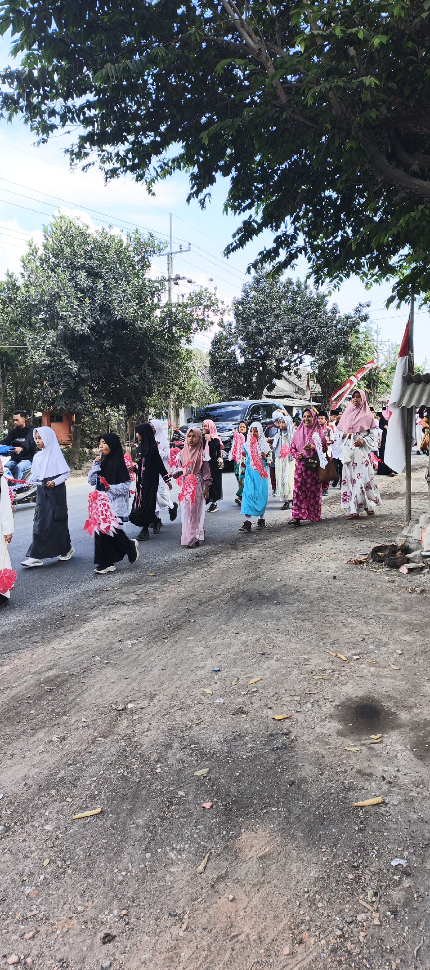 In Grenden, Indonesia, a group of young girls and women are walking down a dirt road. They are dressed in traditional Islamic attire, including hijabs and modest dresses. Some of the girls are carrying festive decorations, possibly for a parade or celebration. In the background, trees line the road, and a black car is parked. The sky is partly cloudy. The ground is covered with dirt and some scattered debris. A red and white flag, possibly the Indonesian flag, is visible on the right side of the image.