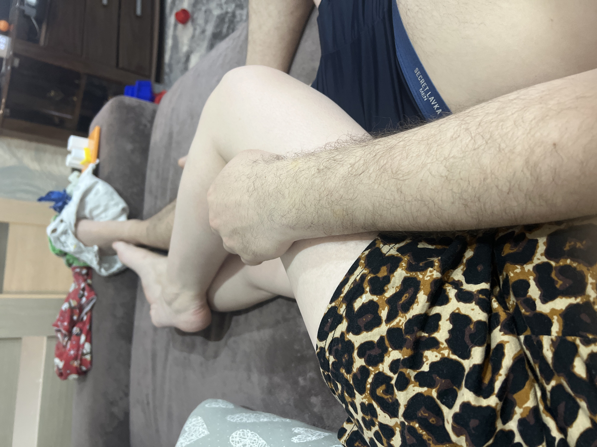 The image shows a close-up, interior view of two individuals, likely on a couch. One person is wearing dark shorts with the waistband of underwear reading "SECRET LAVKA MEN" visible. The other person is wearing shorts with a leopard print pattern. There's a gray couch visible, and to the left, a shelf containing various objects and fabrics. No information suggests the image was taken in the city of Noviy, Russia.