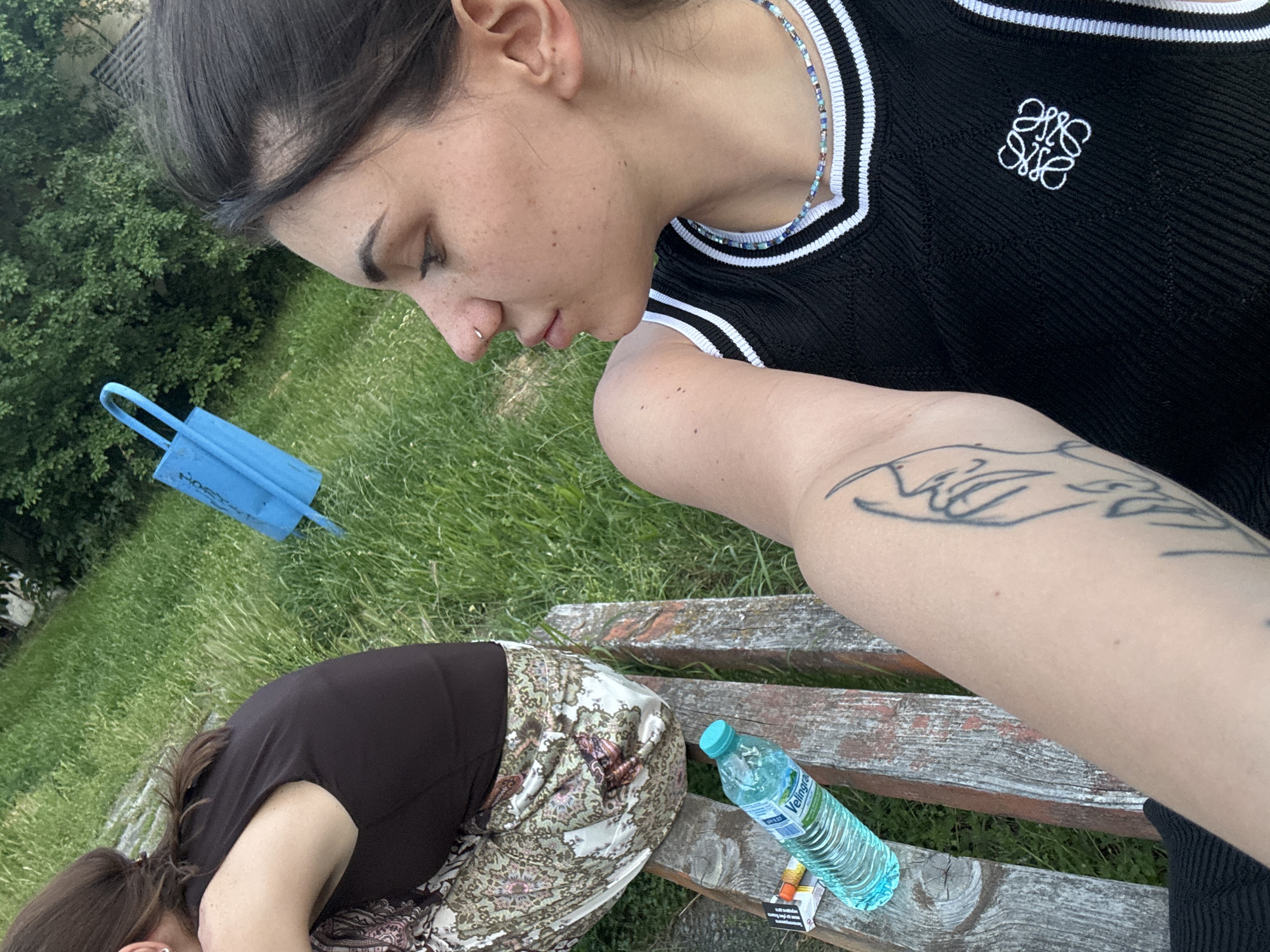 In Sofia, Bulgaria, two individuals are seated on a wooden bench outdoors on a grassy area. One individual, in the foreground, is facing away from the camera and has a tattoo on their arm. They are wearing a black sleeveless top with white trim and a beaded necklace. In the background, another individual is seated, wearing a dark top and patterned skirt. A bright blue bag is visible on the grass behind them. On the bench next to the foreground individual, a bottle of water and a pack of cigarettes are present. The environment appears to be a park or outdoor public space with trees and lush green grass.