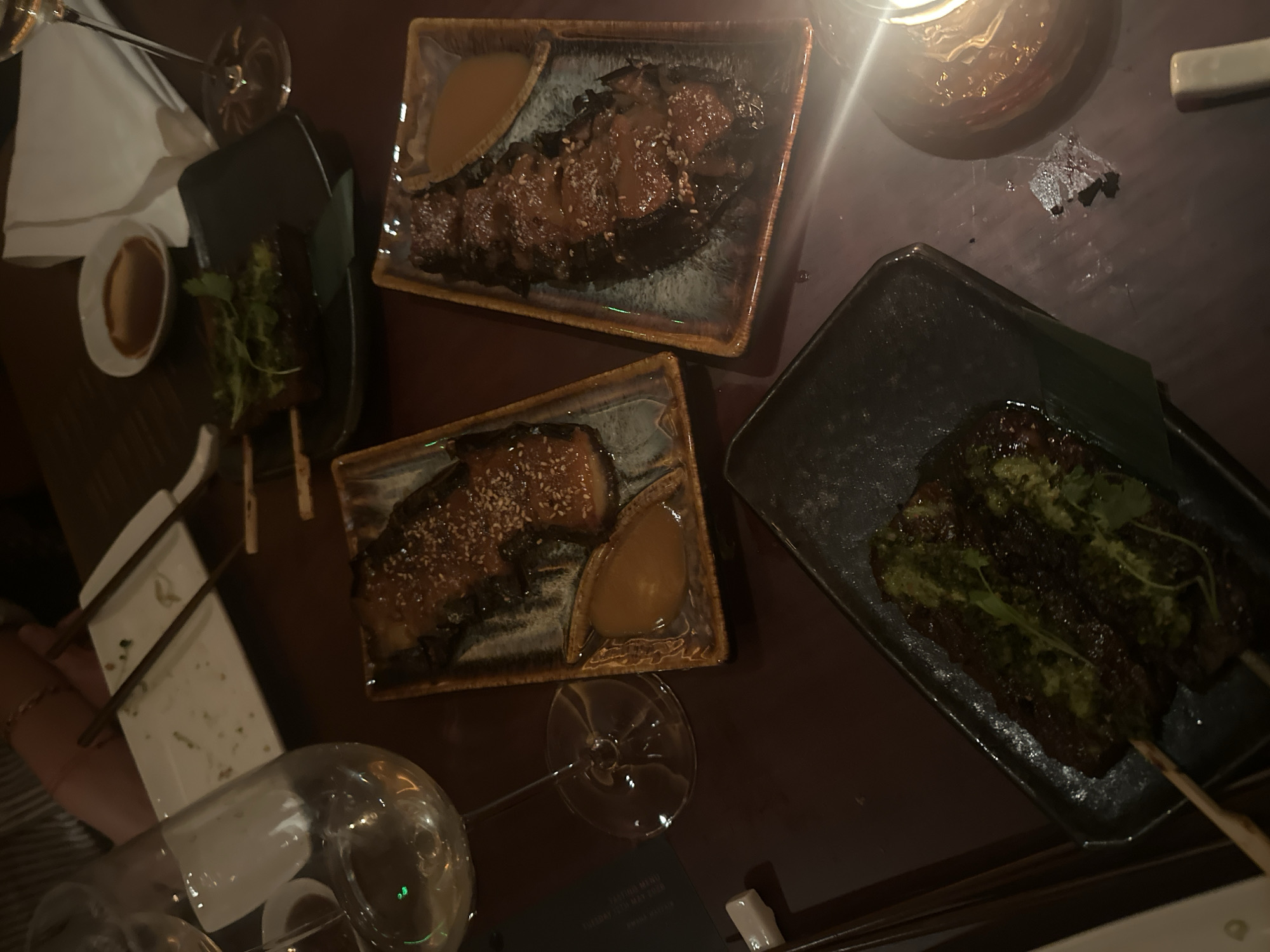 The image depicts an overhead shot of a table with various dishes, likely in a restaurant. The table has a dark surface and features several plates containing different food items.
On the table, there's a rectangular plate with dark-colored meat drizzled with a brown sauce and sesame seeds, along with a small dish of dipping sauce. Another similar plate shows a smaller portion of meat with a similar sauce. A third plate contains grilled meat skewers topped with a green herb sauce. There is also a small bowl of what appears to be dipping sauce near the edge of the table.
Various pieces of cutlery, including chopsticks, are scattered around the table. Additionally, there are wine glasses, one nearly empty and the other partially filled.
The environment appears dimly lit, with a light source visible in the upper right corner casting a glow. There are also other dining accessories, such as plates and possibly a menu, present on the table.
The image provides no information about the city of London, or the United Kingdom, so it is impossible to give the exact location of the photo.