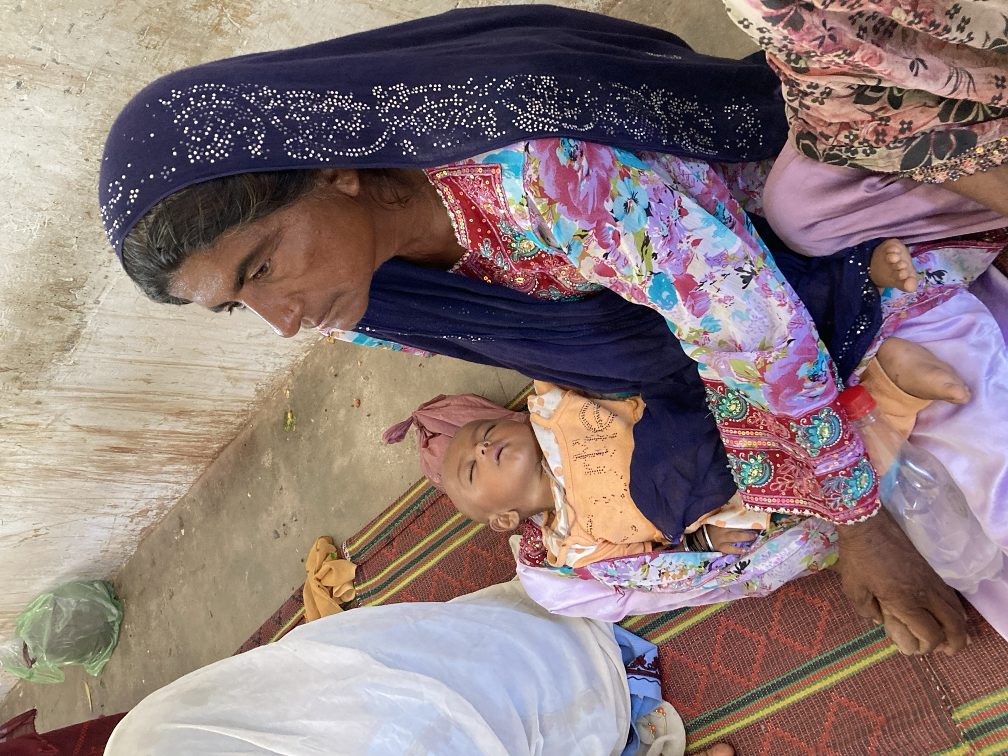 In Goth Ali Marād, Pakistan, an adult woman wearing a navy blue, beaded head covering and a floral patterned garment looks down at an infant in her arms. The infant, wearing an orange garment, is wrapped in lavender-colored fabric and appears to be asleep. The woman sits on a striped mat placed on a bare surface. Other fabrics and items, including a plastic bag and a water bottle, are scattered around the scene.