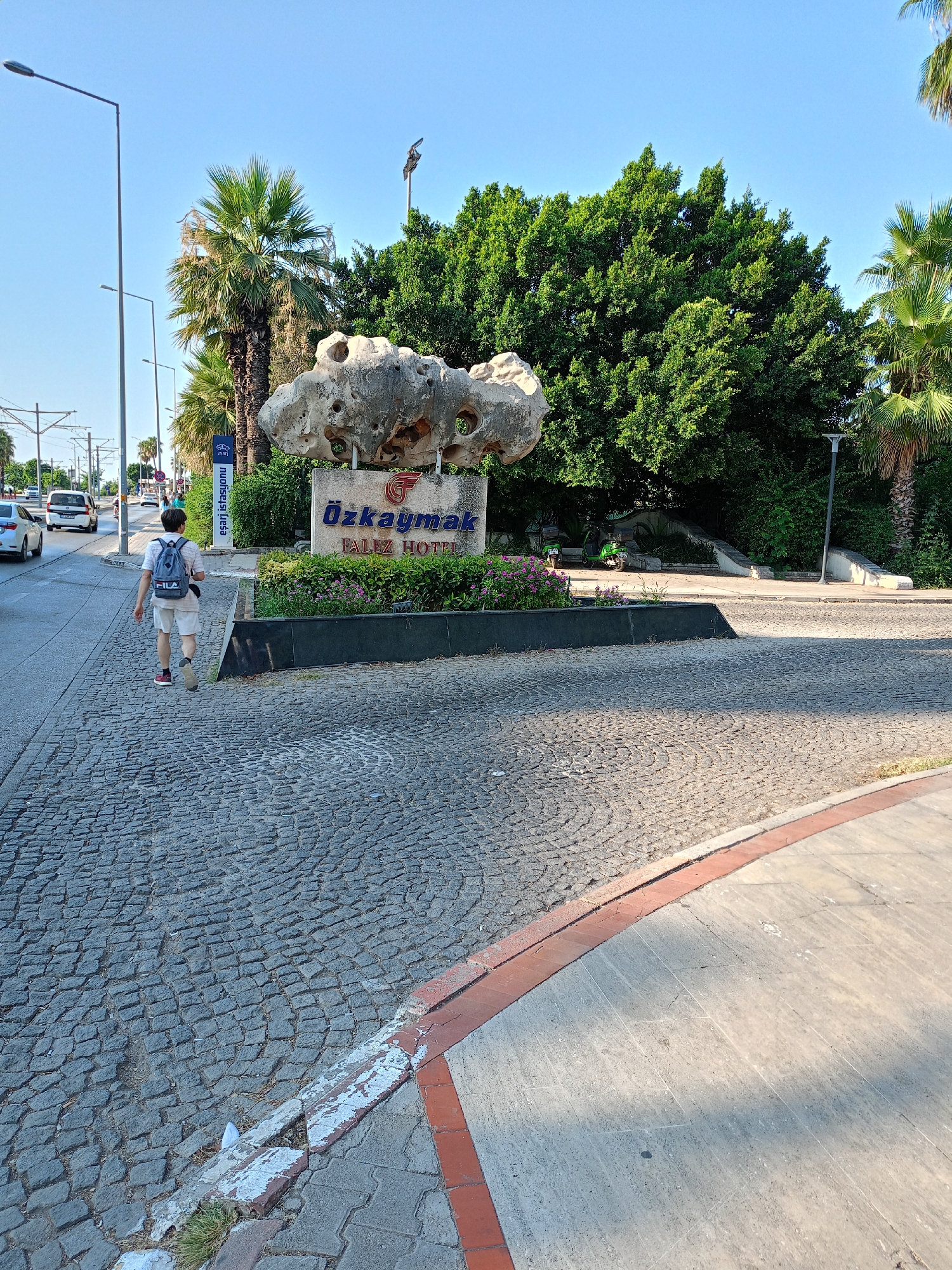 A young male with a backpack walks away from the camera down a cobblestone sidewalk on a sunny day in Antalya, Turkey. He is wearing a white t-shirt, shorts, and red sneakers. He walks past a large sign for "Özkaymak Falez Hotel" which is situated in front of lush green trees and a peculiar rock formation with holes. To his left, there is a street with cars and a bus. The sky is clear and blue. The environment suggests a warm climate, typical of Antalya, a popular tourist destination on Turkey's Mediterranean coast.