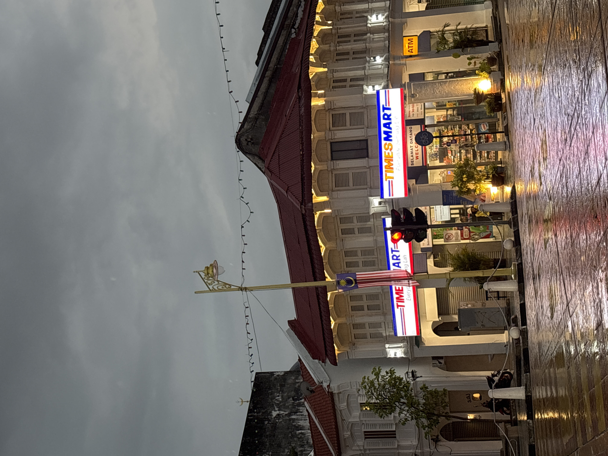 The image shows a street scene in George Town, Malaysia, on a cloudy day, possibly after rain. In the foreground, a wet, tiled street reflects the lights from the buildings. A traffic light is visible, with the red light illuminated. A street lamp with an ornate gold-colored pole stands to the left of a building facade. The building is a shopfront with a red tiled roof and white shuttered windows. A prominent sign for "TIMESMART" is displayed, along with the Malaysian flag banner. An ATM sign is visible to the right. In front of the shop, potted plants are arranged. To the far left, a motorcycle is parked. The architecture suggests a historic street in the city.