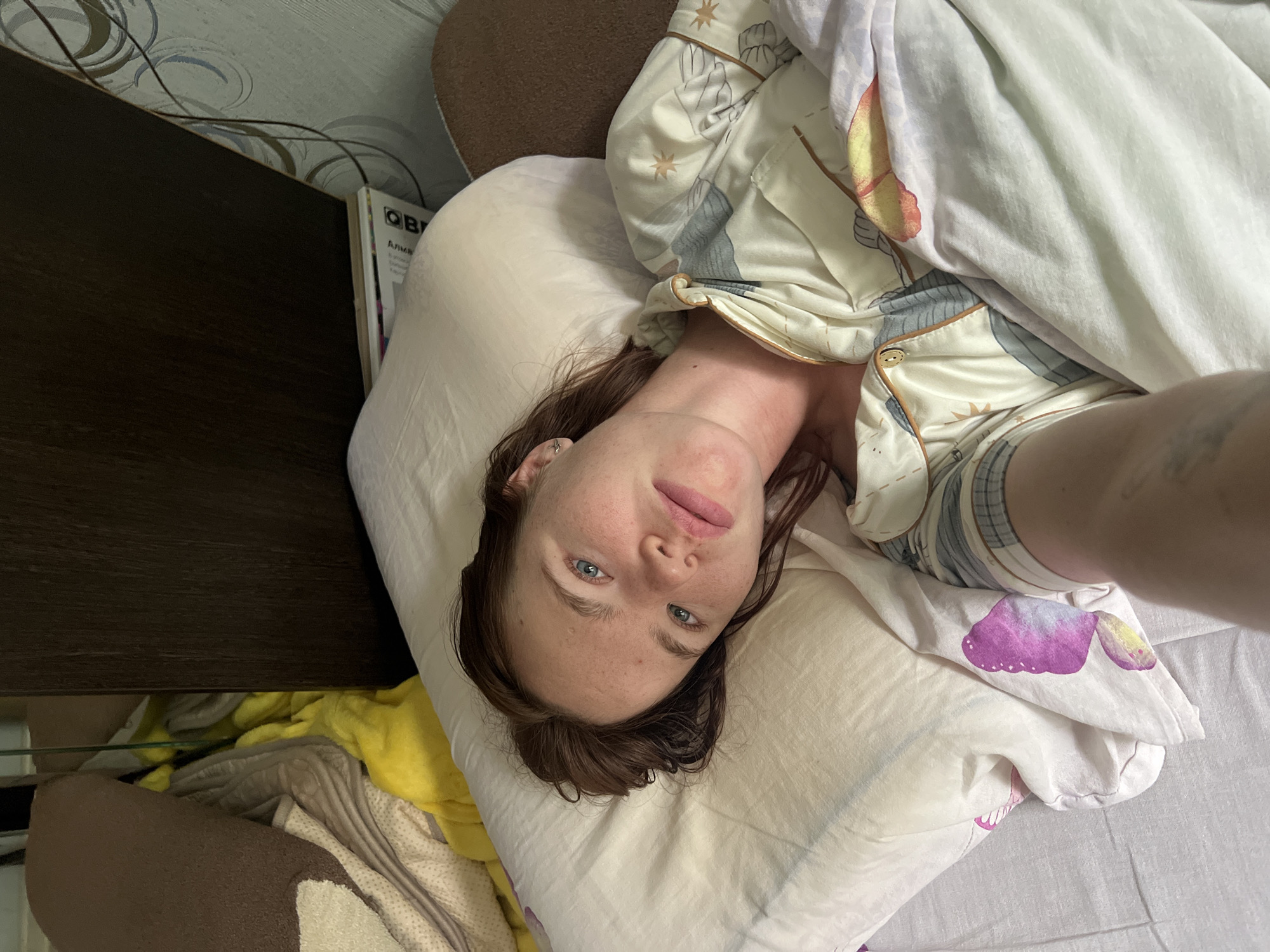 A young woman with brown hair and blue eyes lies in bed, propped up on a pillow and covered by a blanket. She is wearing pajamas with a celestial print. The bed is positioned near a dark wooden headboard, and to the left, a pile of blankets and pillows, including a bright yellow one, is visible. A small book with text in Cyrillic is partially visible on the bedside table behind the pillow. The room appears to be a bedroom in Chaikovsky, Russia.