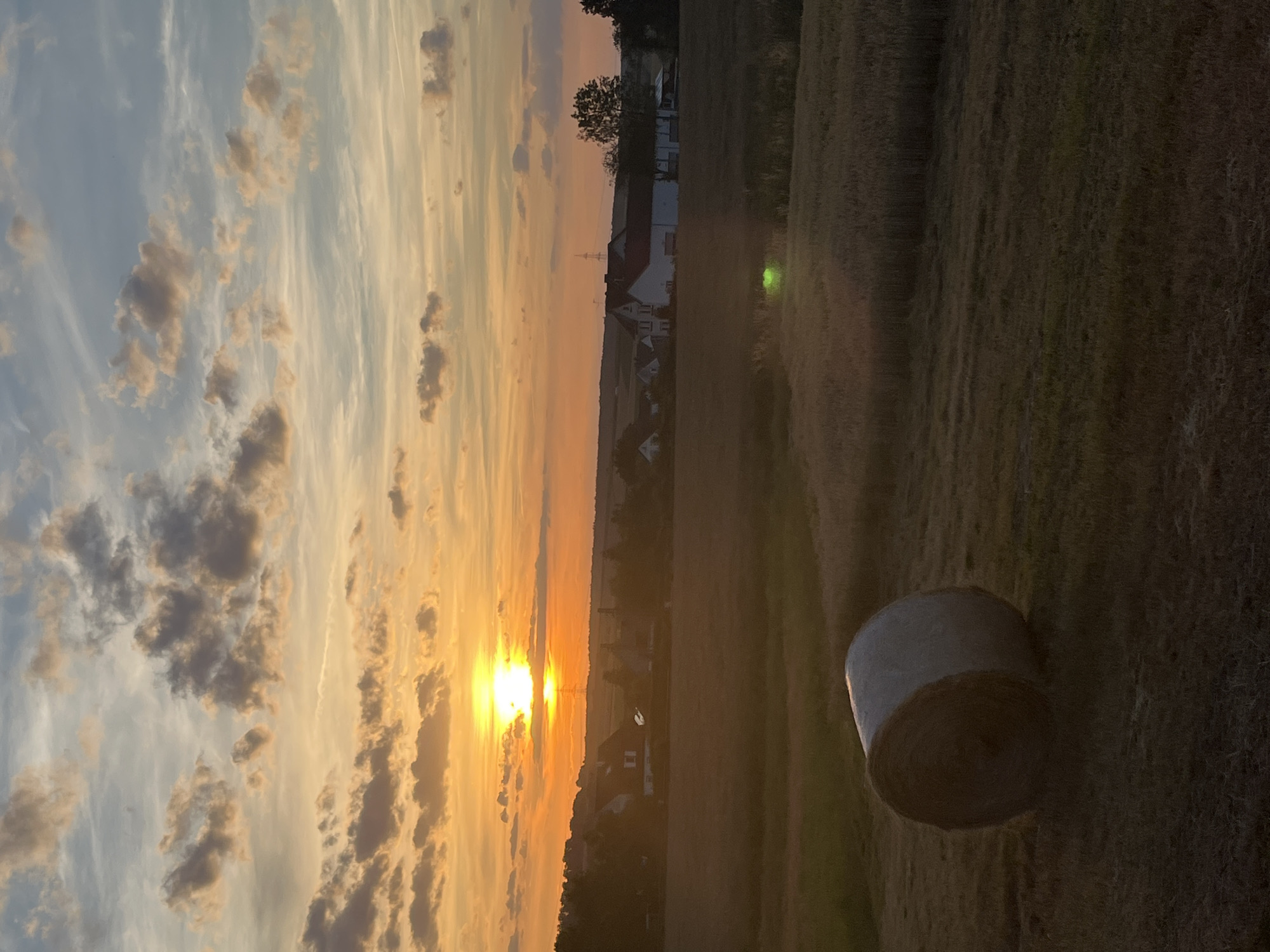 This image depicts a rural landscape during sunset, likely in Neustadt, Germany, given the architectural style of the buildings. The sun is low on the horizon, casting a warm, orange glow across the sky, with scattered clouds tinged with yellow and pink. In the foreground, a large, round bale of hay rests on a harvested field. The middle ground features a cluster of houses with red-tiled roofs, characteristic of rural German architecture, surrounded by trees and vegetation. The background shows rolling hills and a vast expanse of sky. The overall scene evokes a sense of tranquility and the end of a day in the countryside.