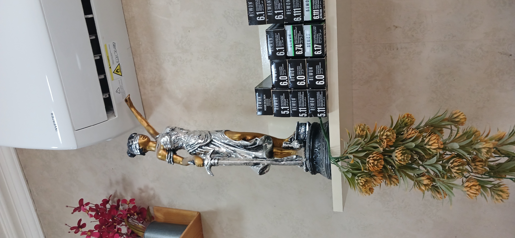 In an indoor setting, an air conditioner unit is mounted high on a textured wall. Below the air conditioner, a statue of Lady Justice, with her eyes blindfolded and holding a sword and scales, stands on a shelf. To the right of the statue, several boxes of hair dye are stacked. To the left of the statue, a vase of red flowers is visible. In the foreground, a decorative hanging plant with yellow cone-shaped flowers drapes downwards. The image is taken from a slightly low angle, with the shelf extending across the frame. The scene appears to be a retail or salon environment, with the hair dye boxes suggesting a beauty salon. The location is not specified as Ganja City, Azerbaijan.