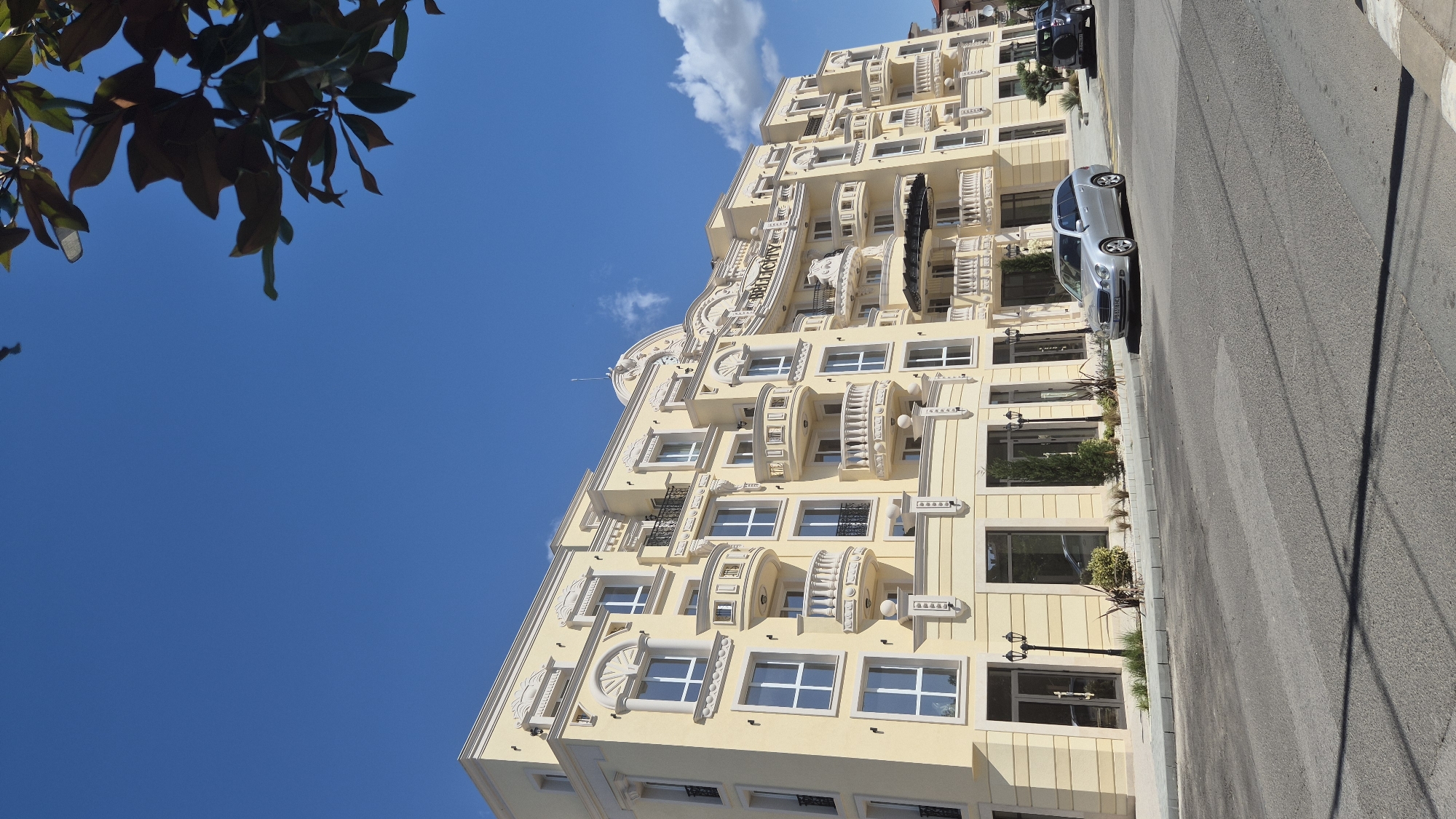 A low-angle, eye-level shot captures the facade of a cream-colored, multi-story building in Ravda, Bulgaria, under a clear blue sky with scattered clouds. The building features ornate architectural details, including decorative window surrounds, balconies with balustrades, and a curved mansard roof with the name "BELLICITY" prominently displayed in gold lettering. Several windows are visible, most with white frames. Greenery, including trees and bushes, is planted along the building's base. A silver sedan and a dark SUV are parked on the street in front of the building. The street is paved, with a crosswalk marking visible. A tree branch with dark green leaves is visible in the upper right corner of the frame. The scene appears to be a sunny day in an urban environment.