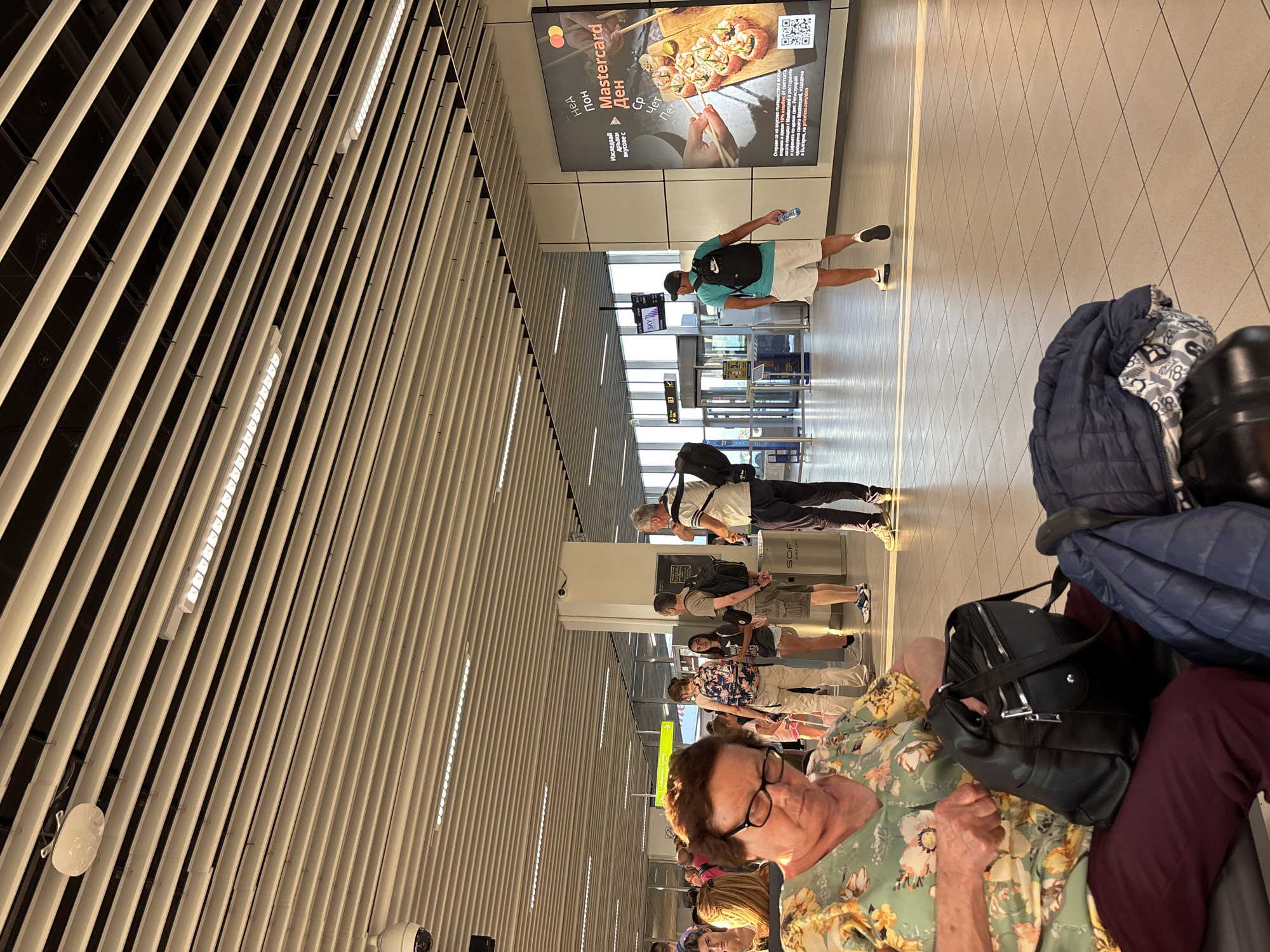 The image depicts a scene inside an airport terminal, likely at Sofia Airport, Bulgaria, indicated by the "SOF AIRPORT" text. The ceiling features a modern design with parallel white slats and integrated lighting. Several people are visible, appearing to be passengers waiting in line or moving through the terminal.
In the foreground, a person wearing glasses and a floral shirt is seated, with luggage beside them. Further down the terminal, other individuals are seen walking and standing. One person, wearing a turquoise t-shirt and white shorts, is walking away from the camera towards a security or boarding gate area, holding a water bottle. Another man, wearing a patterned shirt and shorts, is also visible in the mid-ground.
A large advertisement panel is mounted on the wall, featuring sushi and promoting Mastercard with text that includes "Mastercard", "Ден", and promotional offers like "10% кешбек" (10% cashback). The advertising uses Bulgarian text, further confirming the location. The lighting suggests it is daytime, possibly late morning or afternoon, as natural light is entering the terminal from large windows or open spaces. The overall atmosphere appears to be one of travel and transit, with people engaged in typical airport activities.