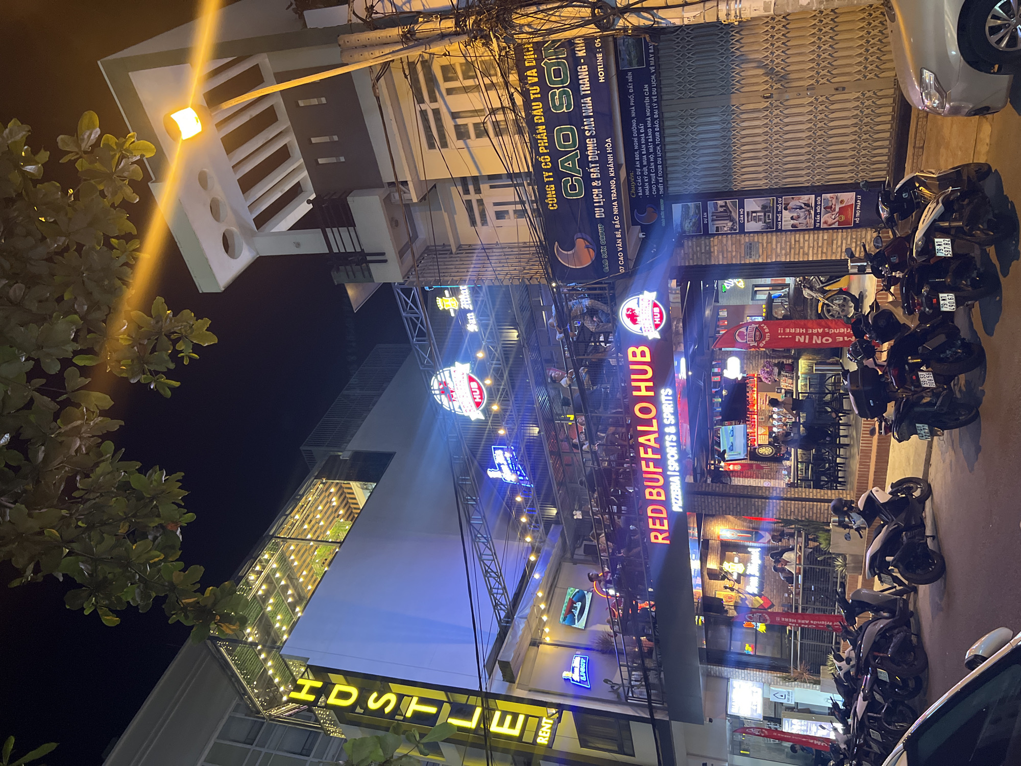At night in Bắc Nha Trang, Vietnam, the scene features "Red Buffalo Hub," a pizzeria and sports bar. The establishment is well-lit with signage in English. Parked motorcycles are visible in the street. The surrounding buildings include the "Hustle Rent" Hostel. Additionally, a sign for the Cao Son Group, a real estate and tourism company, is present, advertising its services, and listing its address as 07 Cao Van Be.